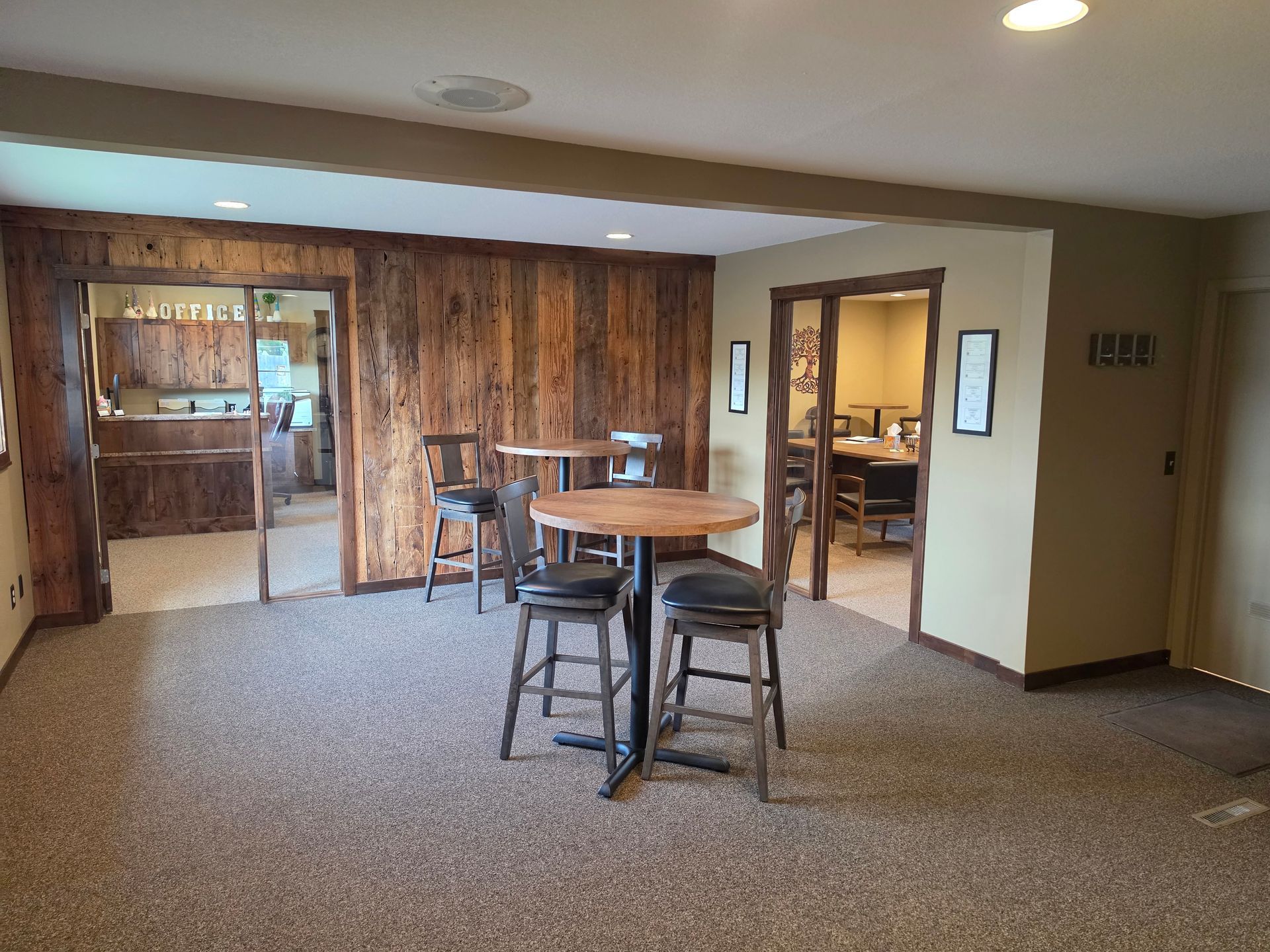 Spacious carpeted room with round tables, stools, wood-paneled wall, and a doorway to another room.