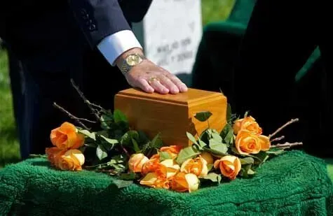 Hand on a small wooden casket surrounded by orange roses on green cloth