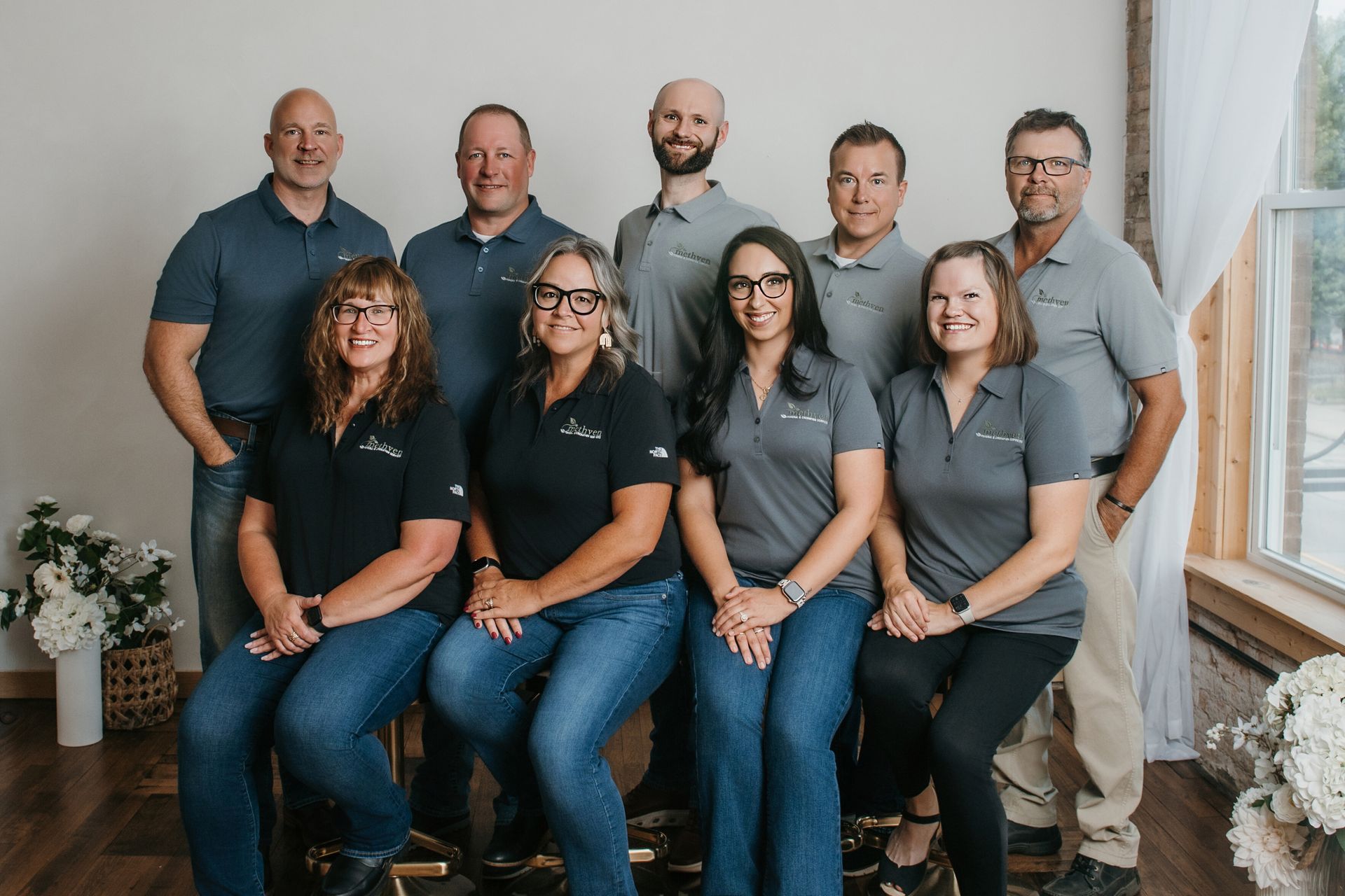Group portrait of eight coworkers in navy shirts posing indoors by a window and white wall
