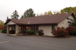 Single-story beige building with brown roof and covered entrance beside a paved parking lot
