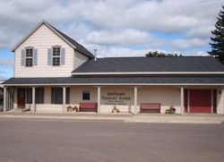 White farmhouse-style building with red doors, porch, and sign in front under a cloudy sky