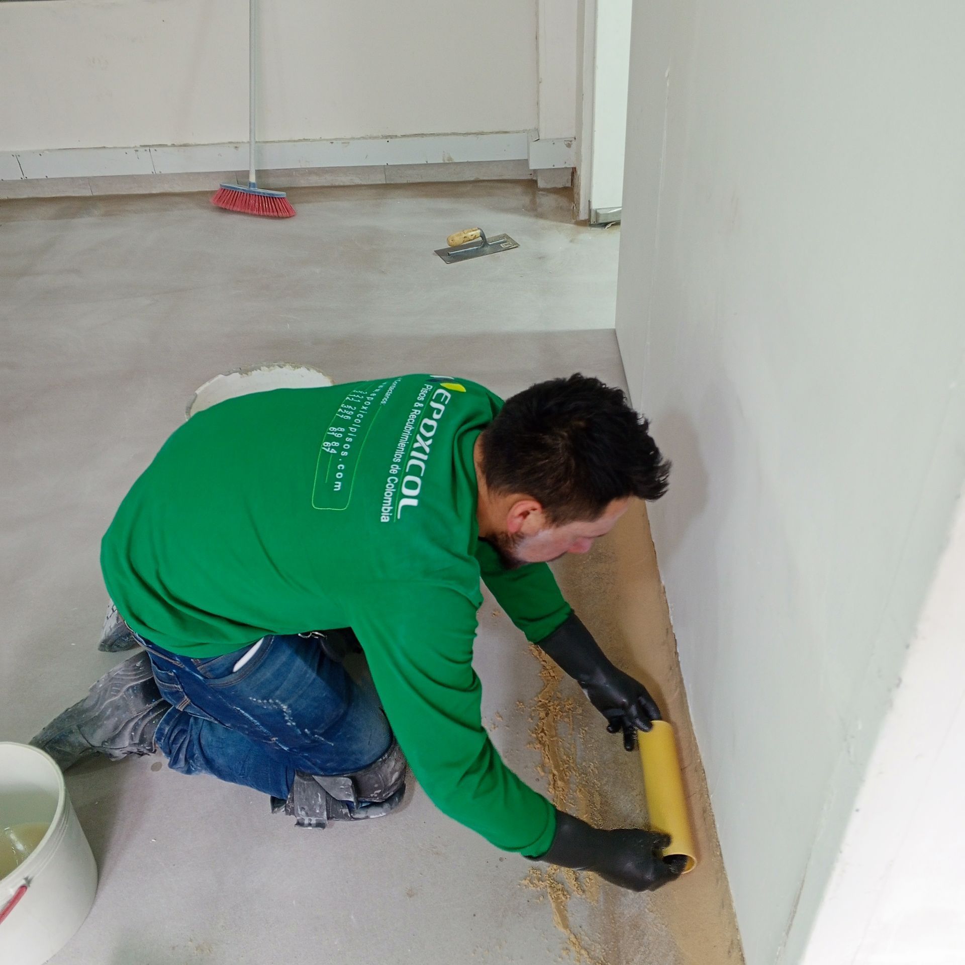 Persona con camisa verde aplicando una sustancia a la pared con un rodillo; ambiente interior.