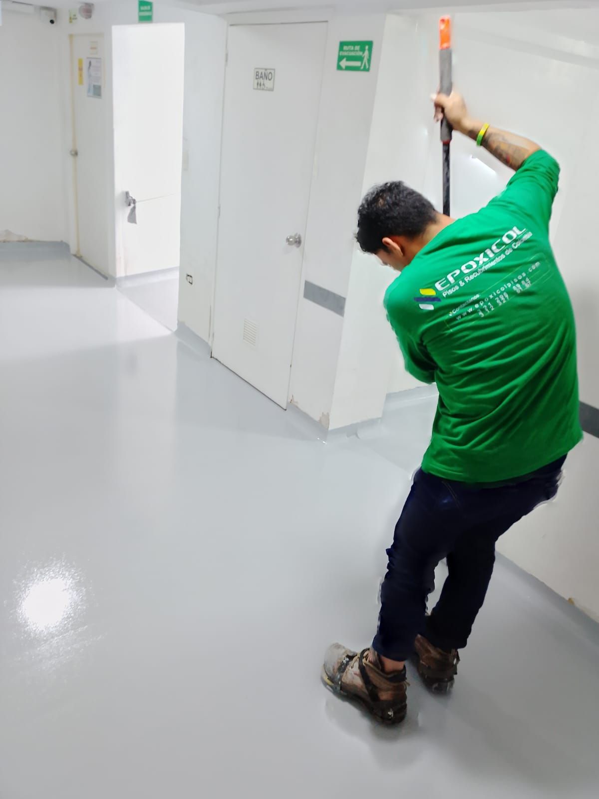 Persona con camisa verde utilizando una herramienta de pértiga en un pasillo blanco con suelo gris.