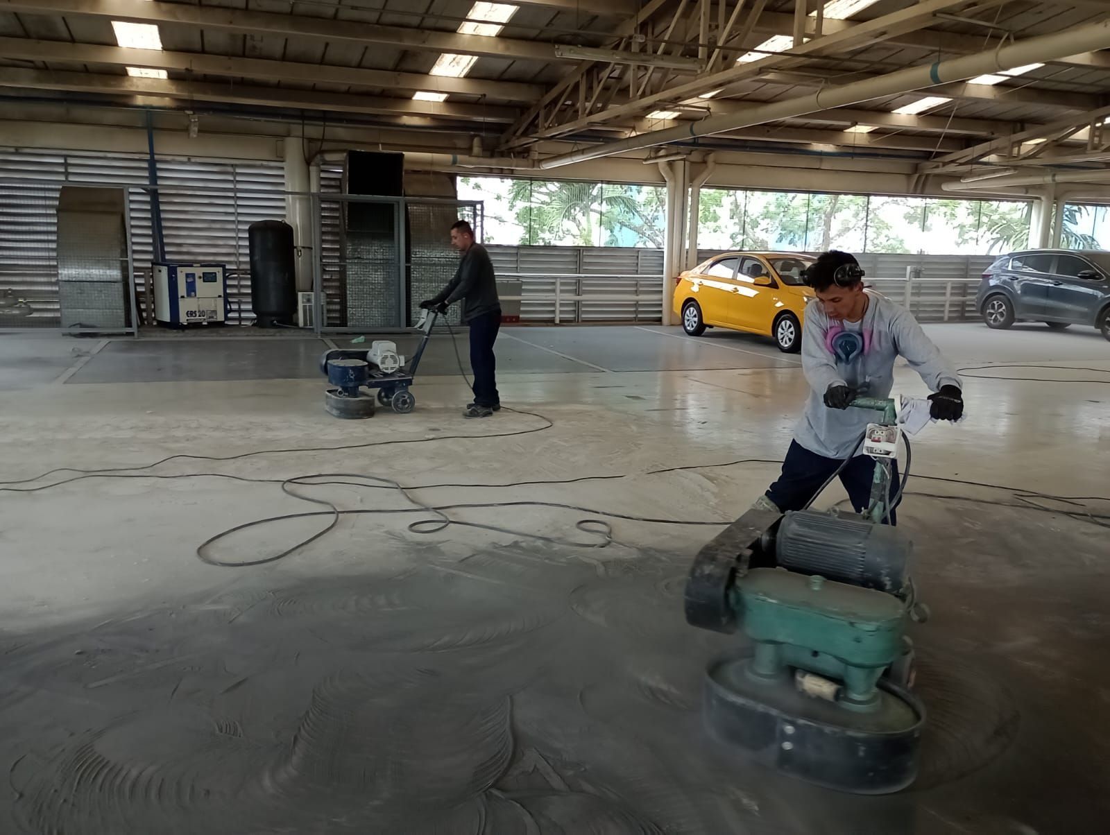 Dos personas puliendo un suelo de hormigón con máquinas industriales en un edificio tipo almacén.