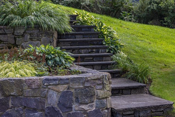 Natural stone retaining wall and stairway surrounded by lush green landscaping, ornamental grasses, sloped backyard hillside.
