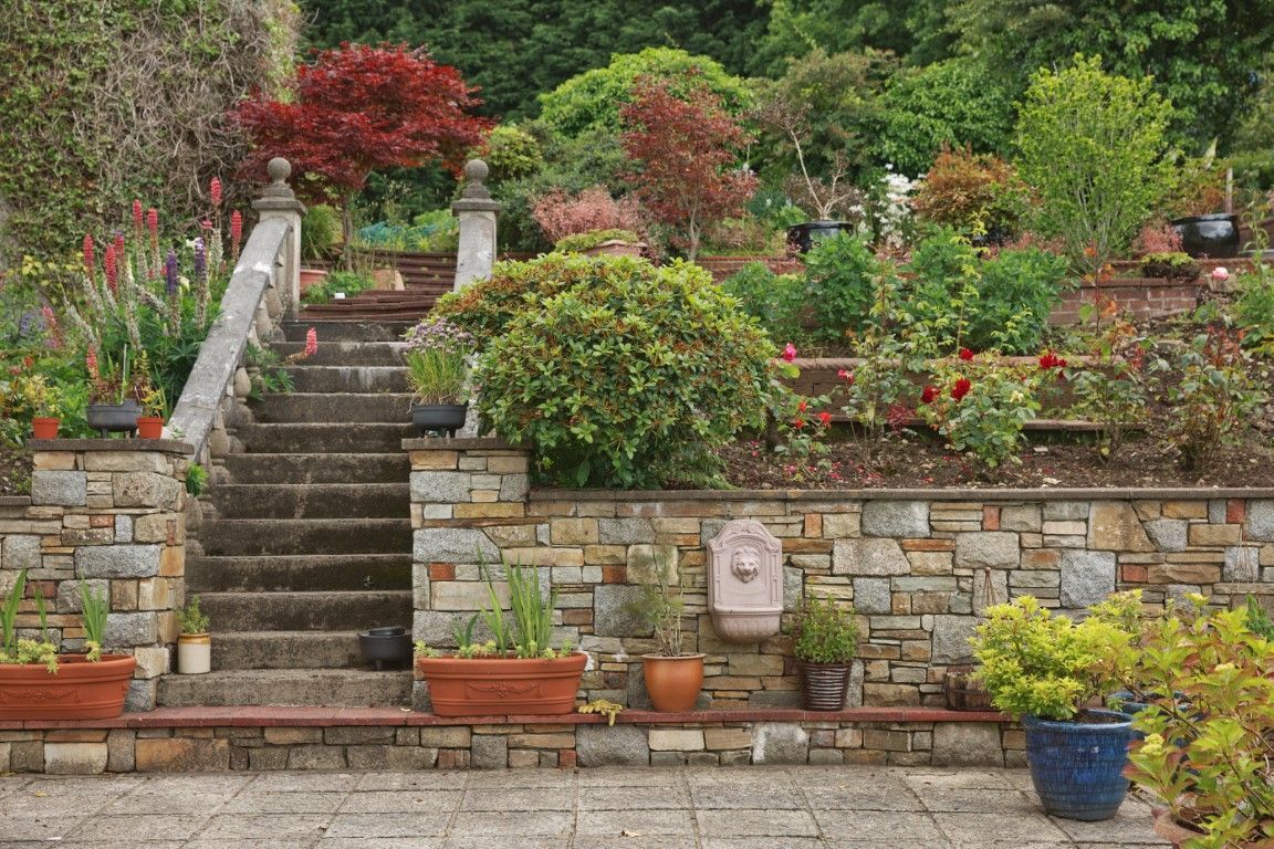 Stone staircase in a tiered garden with colorful plants and flowers; decorative wall fountain.