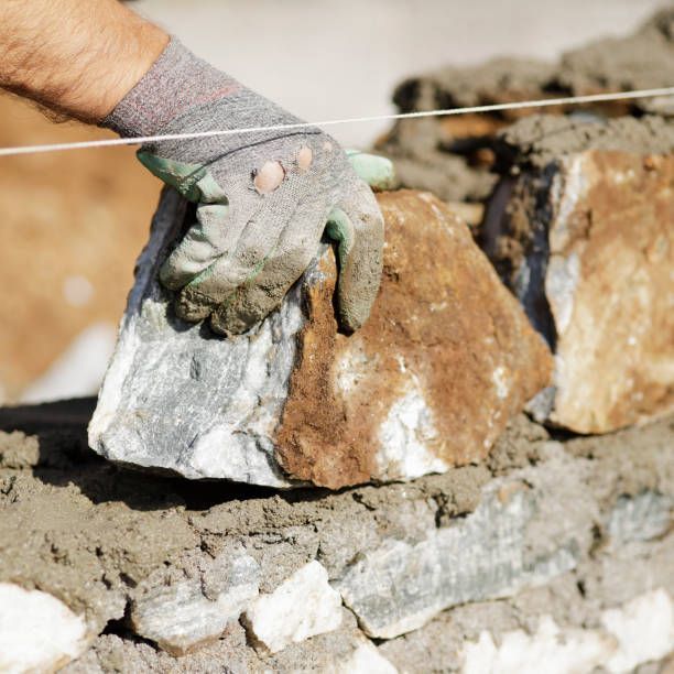 A person places a large, textured stone into a bed of mortar while building a wall along a guide line.