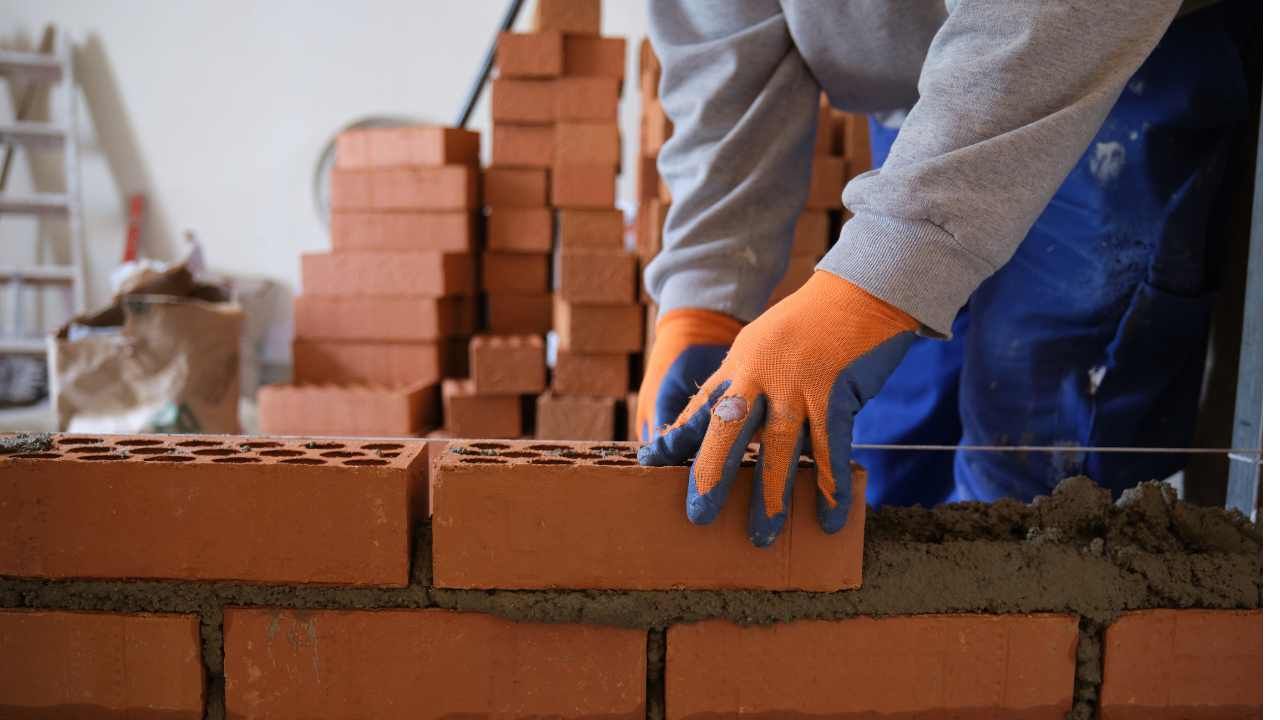 Hands placing red masonry blocks into fresh mortar for a block and retaining wall build