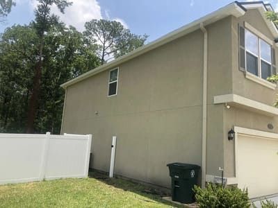 Side view of a beige house with white fence, garage, and green trash bin on a sunny day