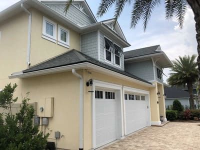 Two-story cream and gray house with white garage doors and a palm tree-lined driveway