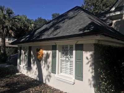 Small white house with a dark shingled roof, window shutters, and a flower basket in the front yard