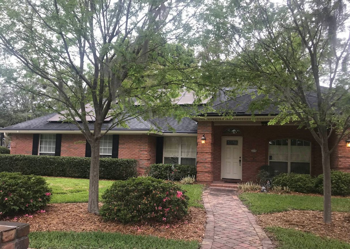 Single-story brick house with a front walkway, trees, and shrubs.