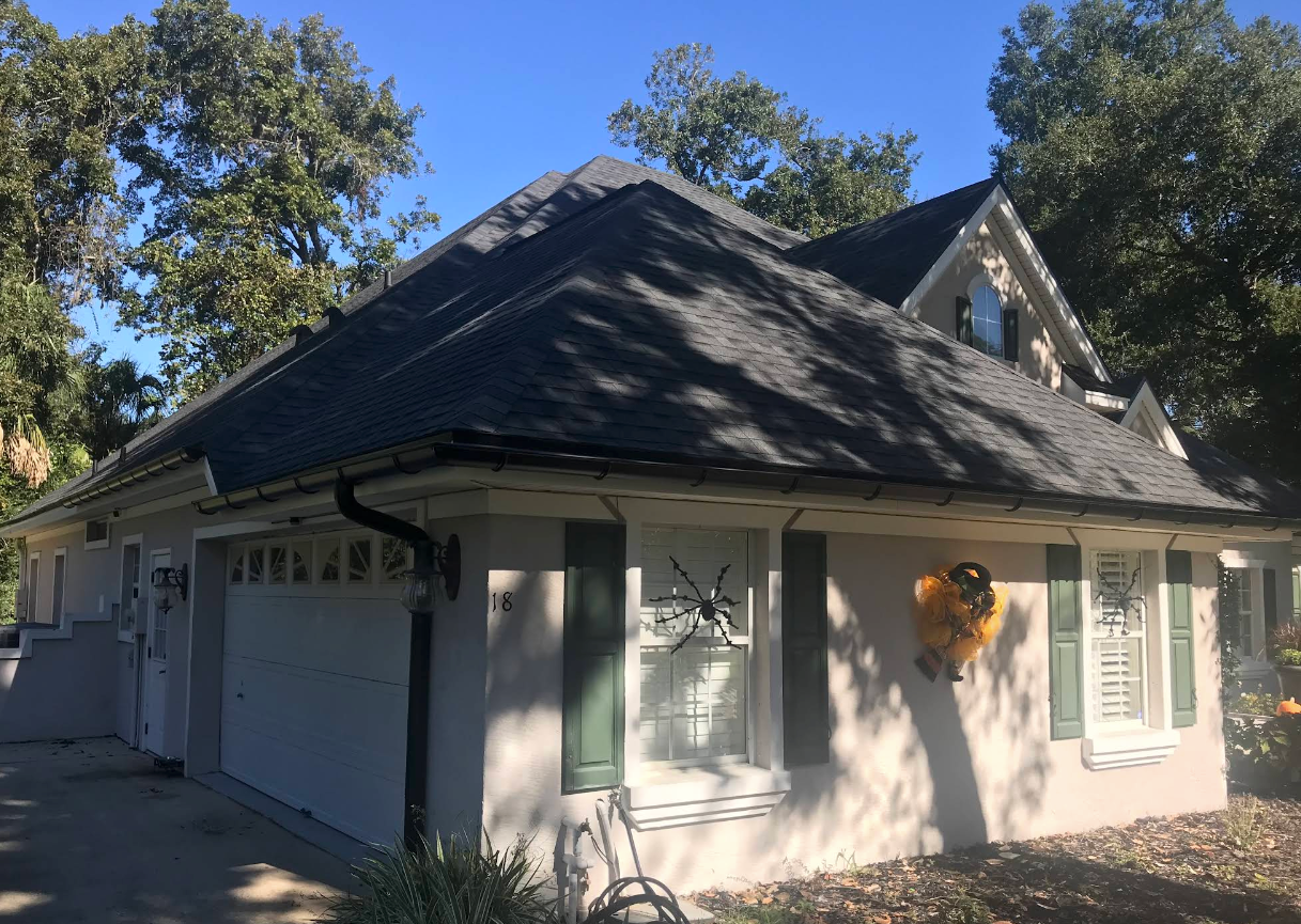 White house with dark roof and green shutters, shown from the front under trees on a sunny day
