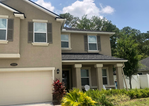 Two-story beige house with white garage, front porch, and green shutters surrounded by landscaping