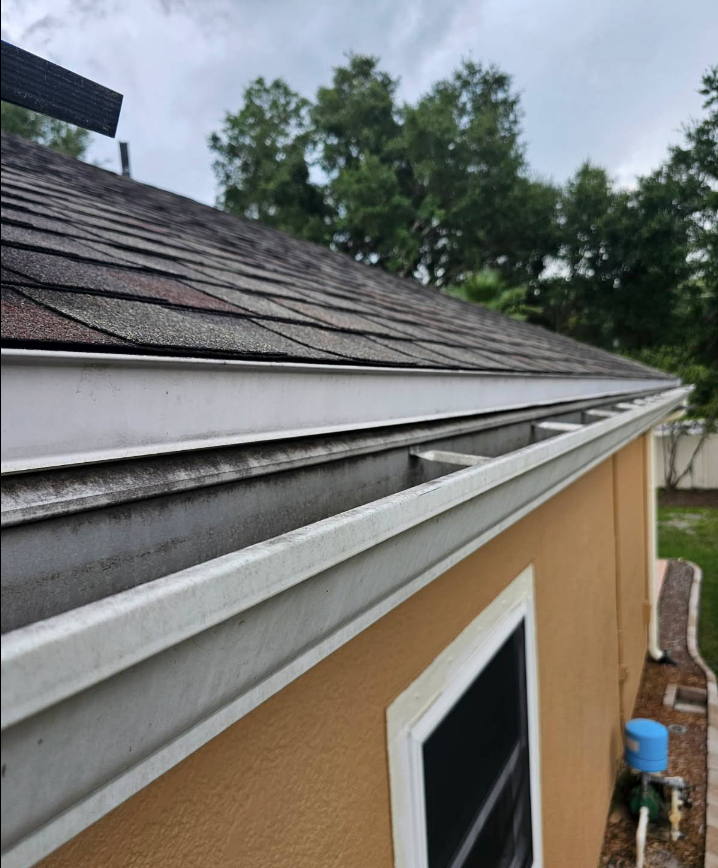 Close-up of a roof edge with white trim and gray shingles above a tan building, with trees in the background