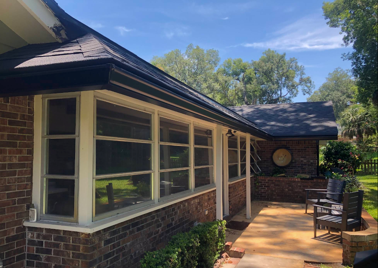 Brick house porch with large windows, dark roof, and patio chairs in a sunny yard