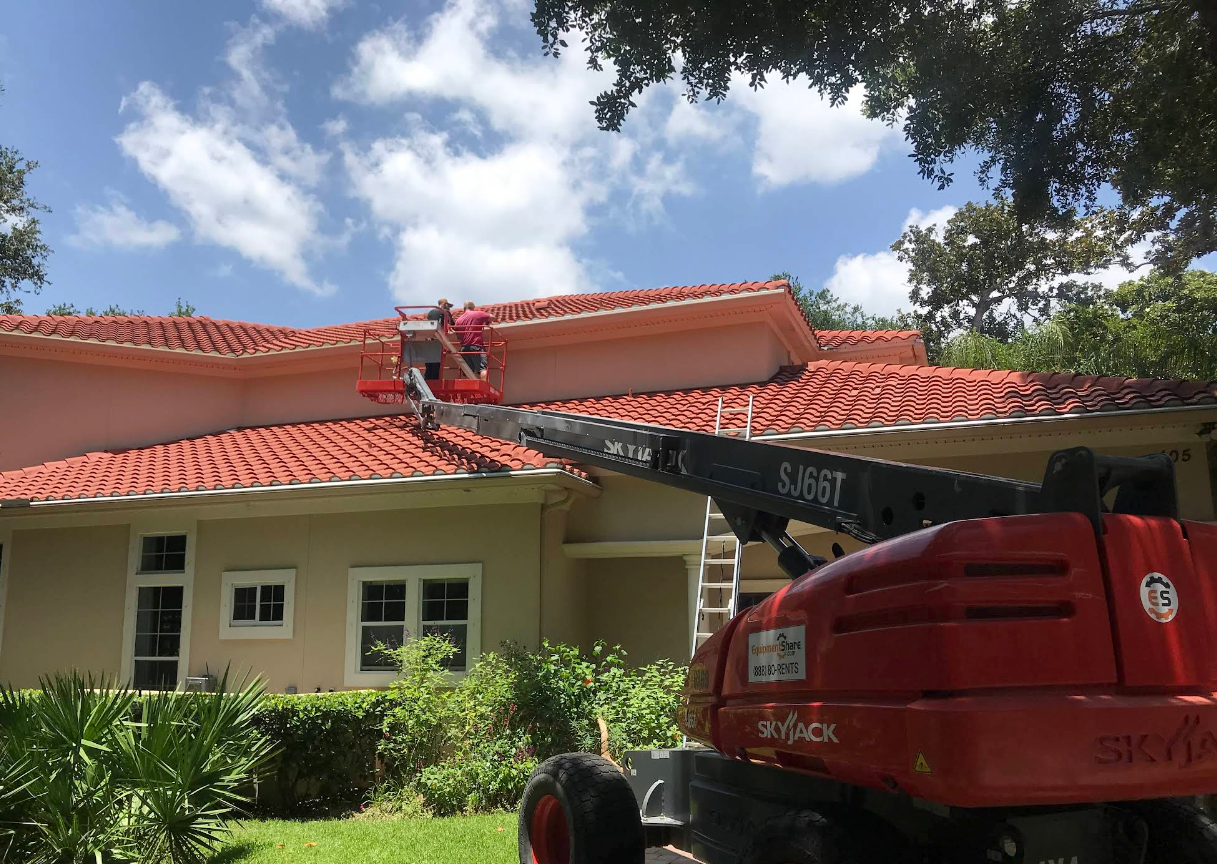Worker on a red boom lift repairing a red-tiled roof on a house under a blue sky.