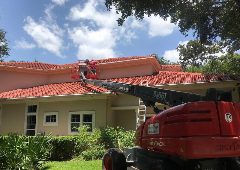 Worker on a red boom lift repairing a red-tiled roof on a house under a blue sky.