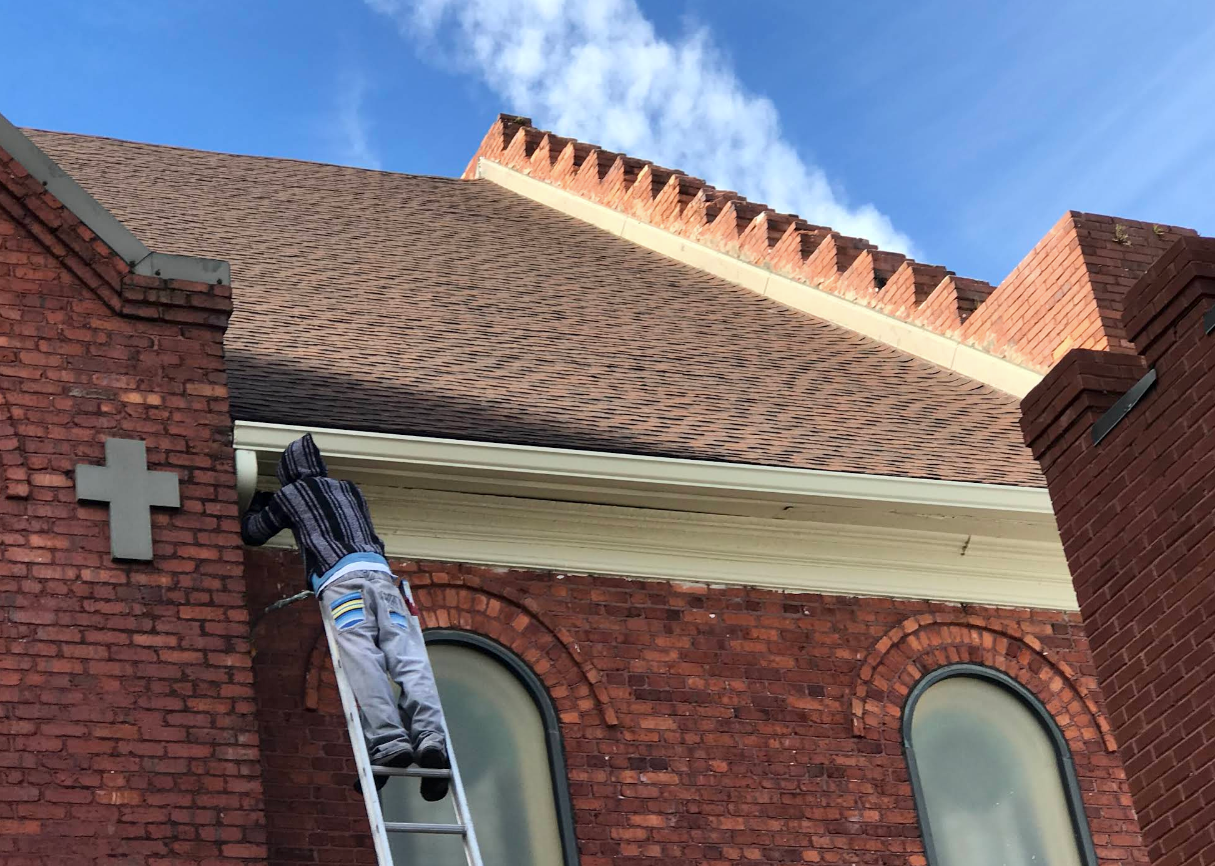 Worker on ladder repairing red brick church roof under blue sky