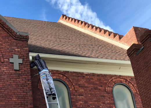Worker on ladder repairing red brick church roof under blue sky