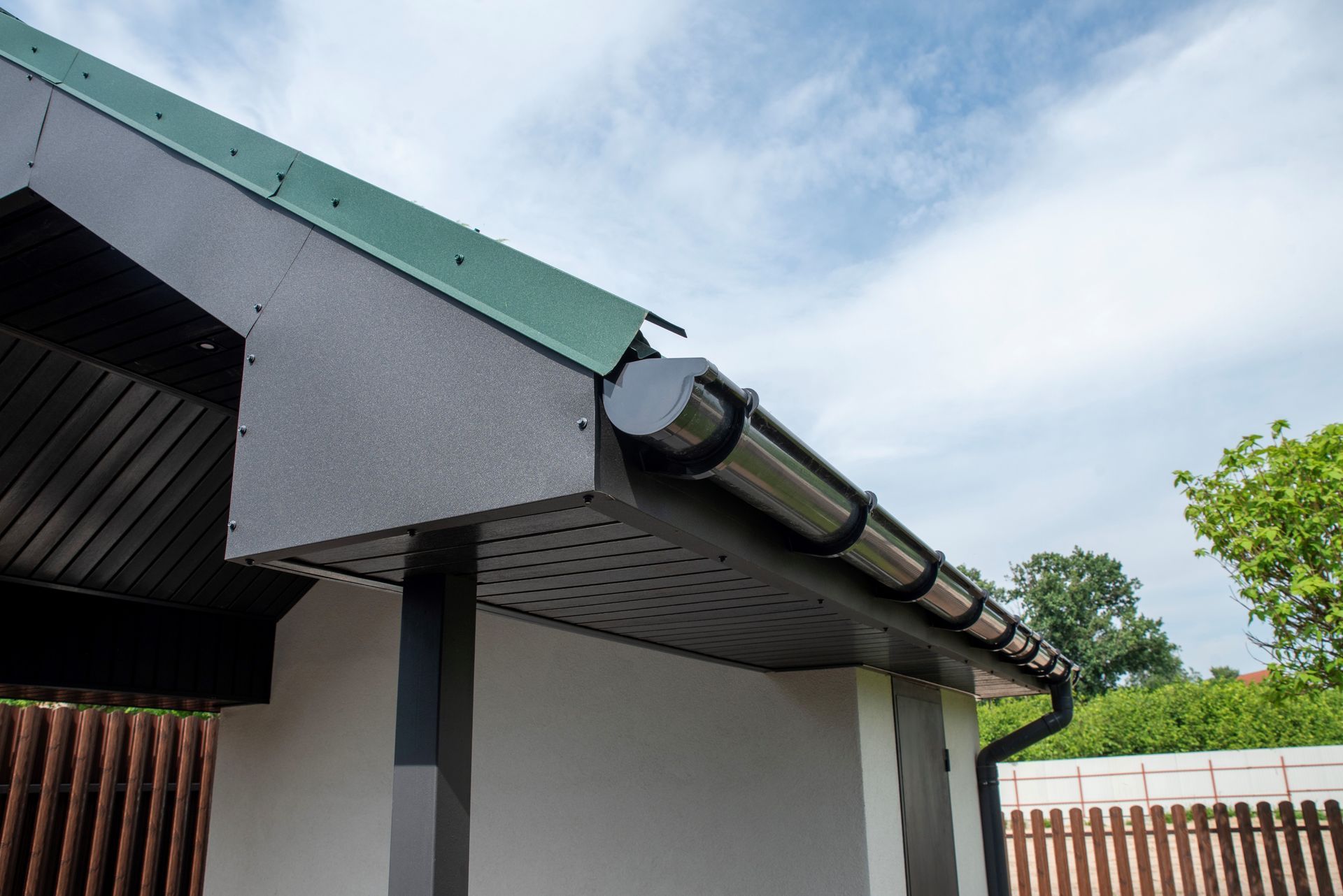 Corner of a house roof with green metal trim, white walls, and a gutter under a cloudy sky