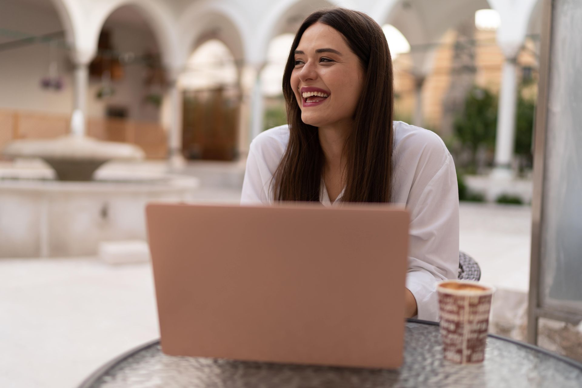 A woman sitting outside on her laptop looking away and smiling.