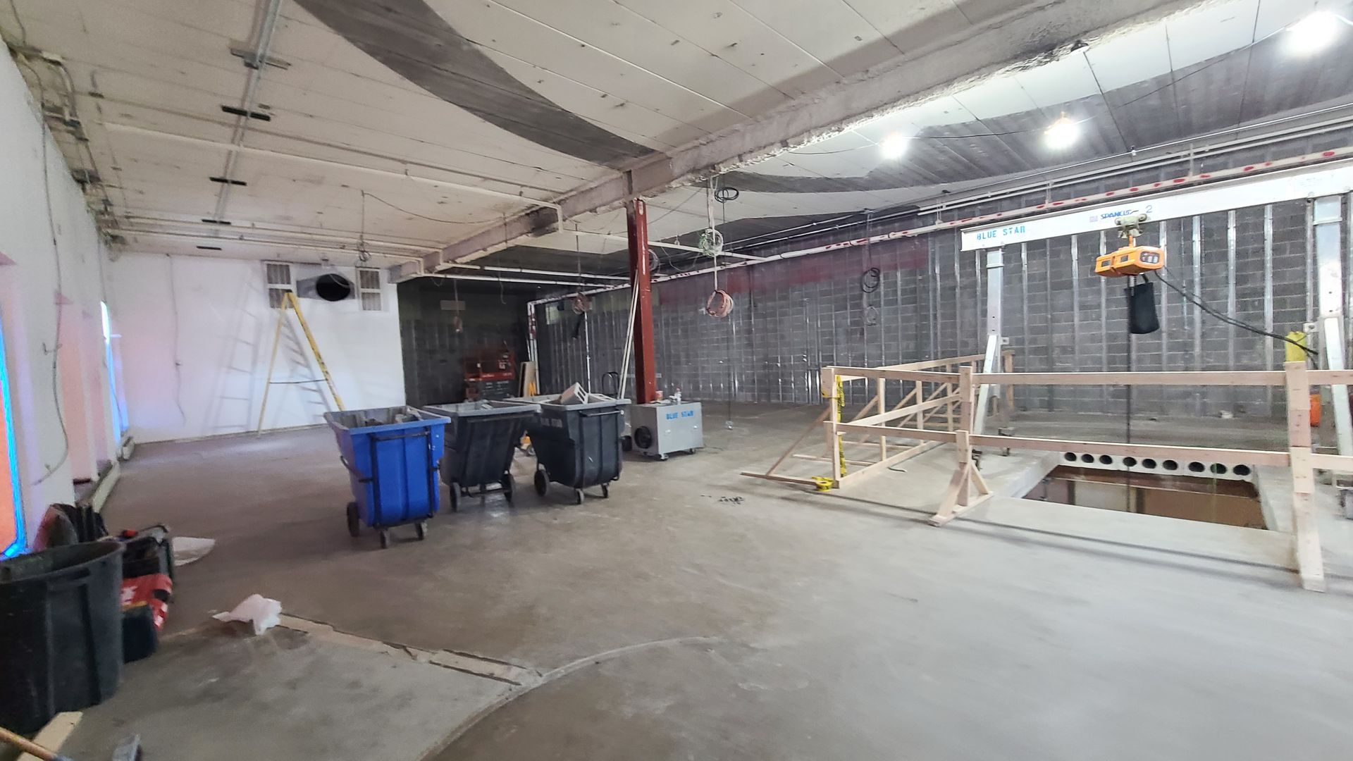 Construction site interior with concrete floors, trash bins, unfinished walls, scaffolding, and overhead lighting.