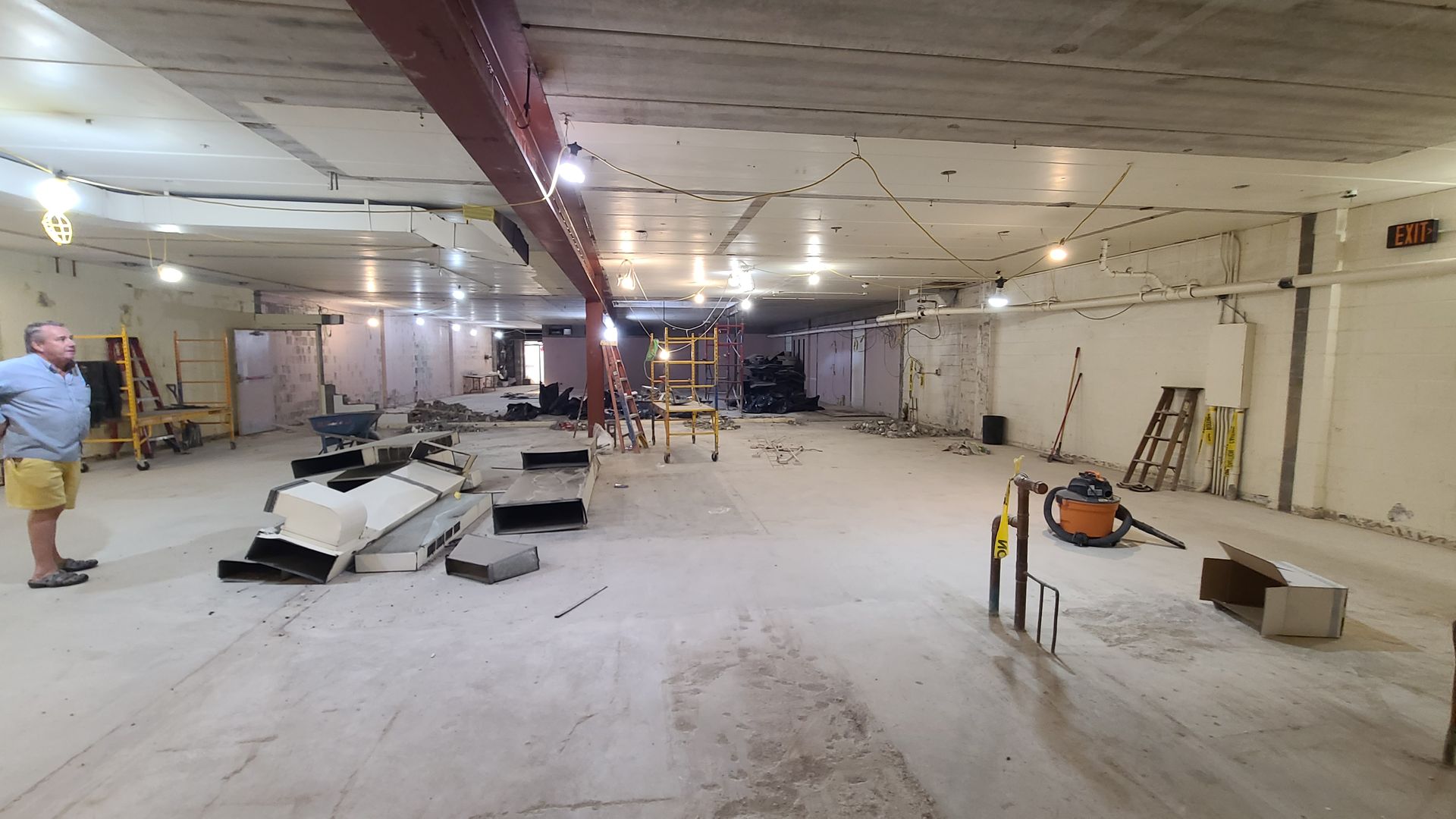 Interior of a construction site: man, debris, tools, open space, overhead lights, exposed ceiling beams.