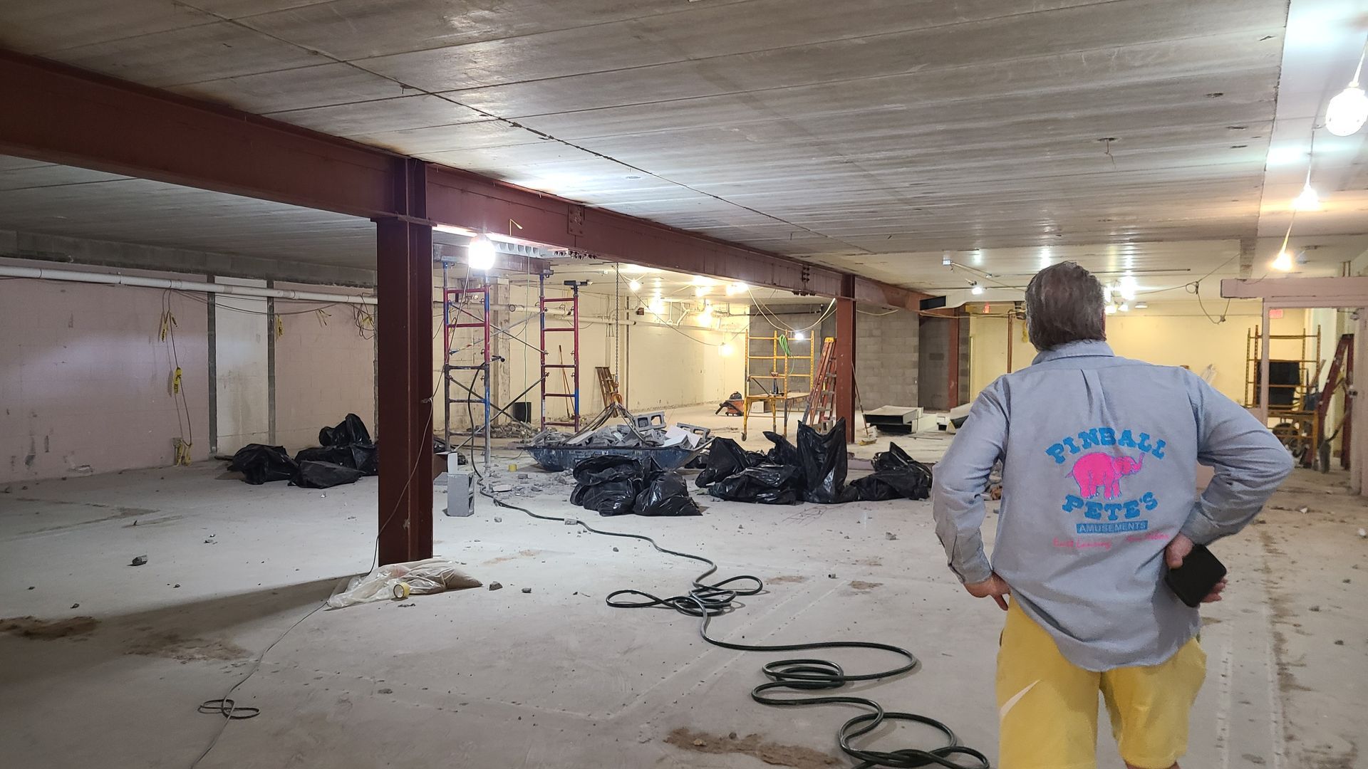 Man in gray shirt surveys a debris-filled basement under construction; steel beams, trash bags.