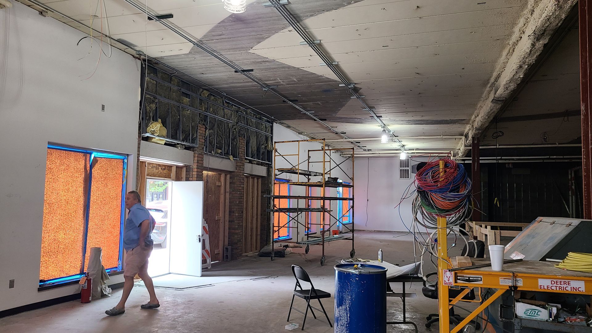 Interior of a space undergoing renovation. Man walks past an open doorway. Exposed wiring, scaffolding, and blue barrels are visible.