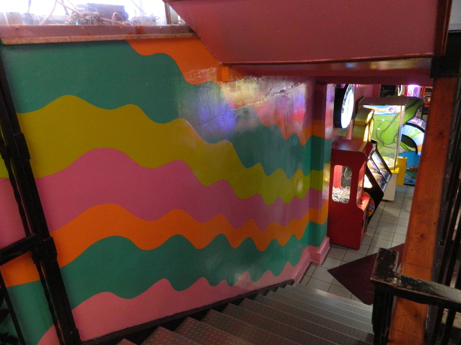 Staircase leading down to a brightly colored arcade room. Wall is painted with wavy bands of green, pink, orange, and yellow.