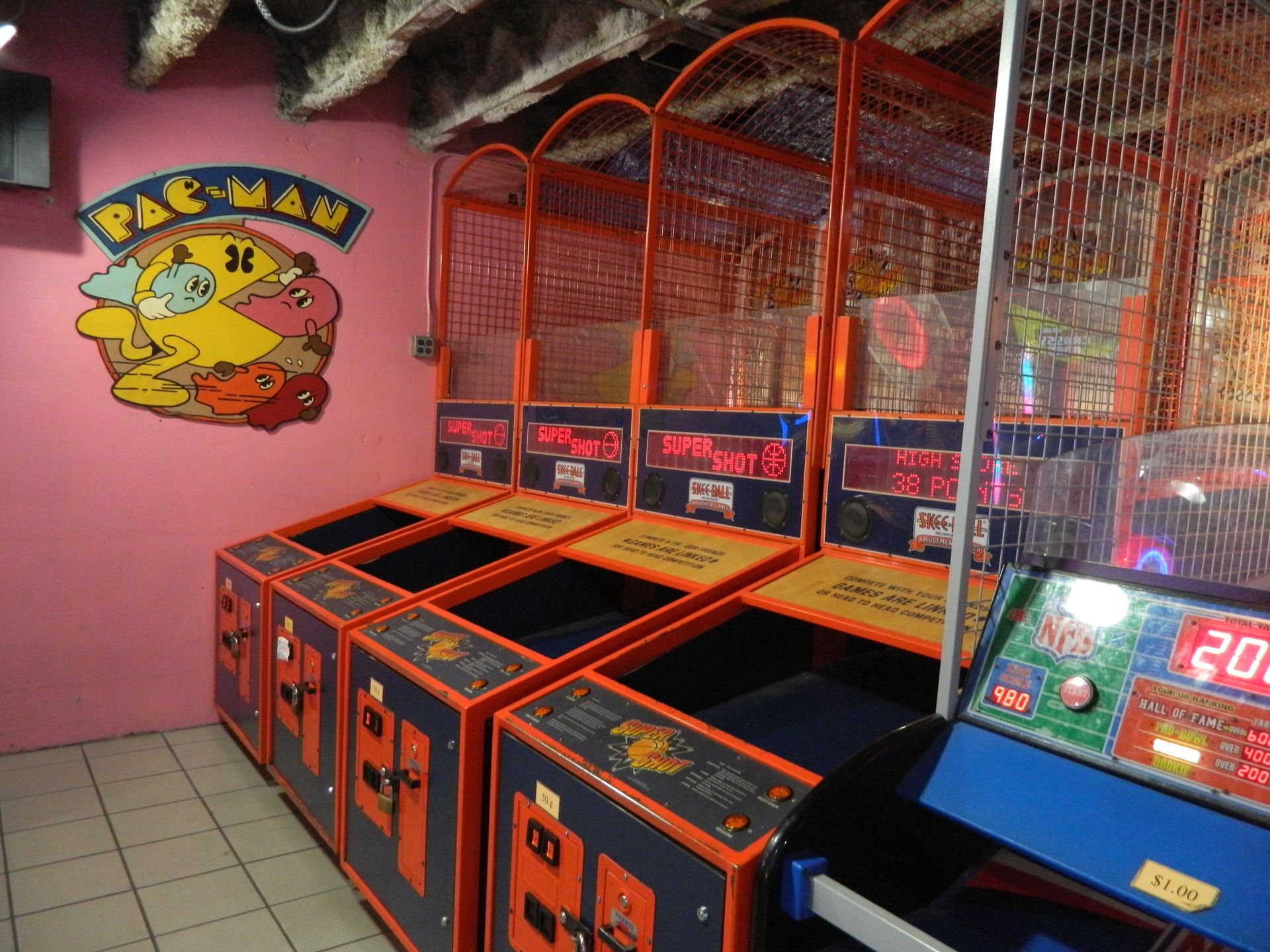 Row of basketball arcade games, orange and blue, with a Pac-Man mural on pink wall.
