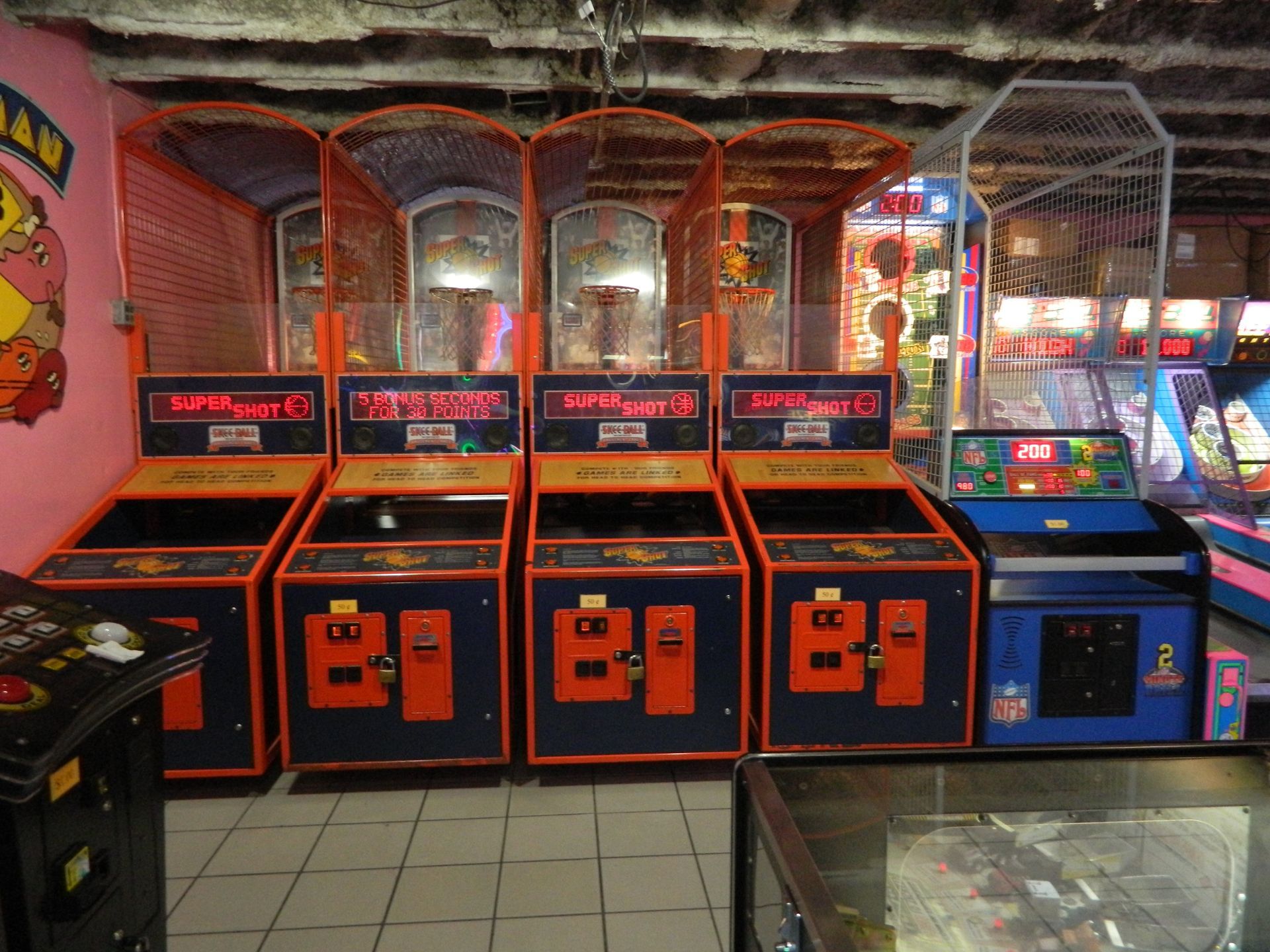 Row of basketball arcade games in an arcade, orange and blue cabinets under metal hoops.
