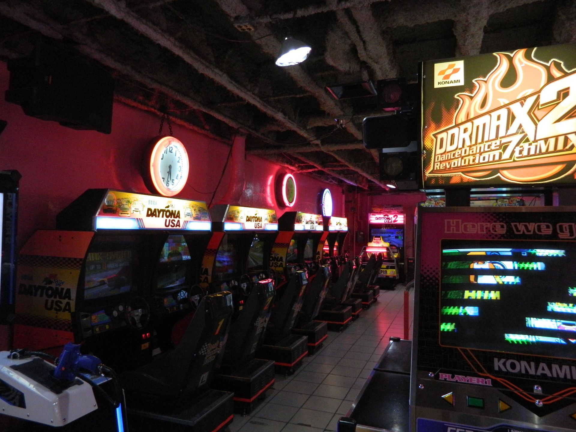 Arcade lined with racing game cabinets, bathed in pink light, overhead pipes visible.