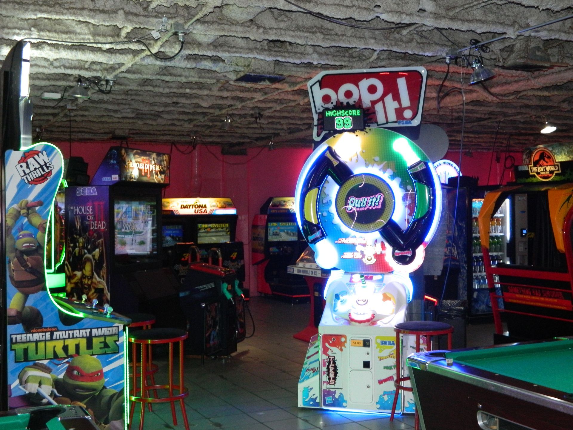 Arcade with various games including Bop It, Teenage Mutant Ninja Turtles, and a pool table. Pink walls, dark ceiling.