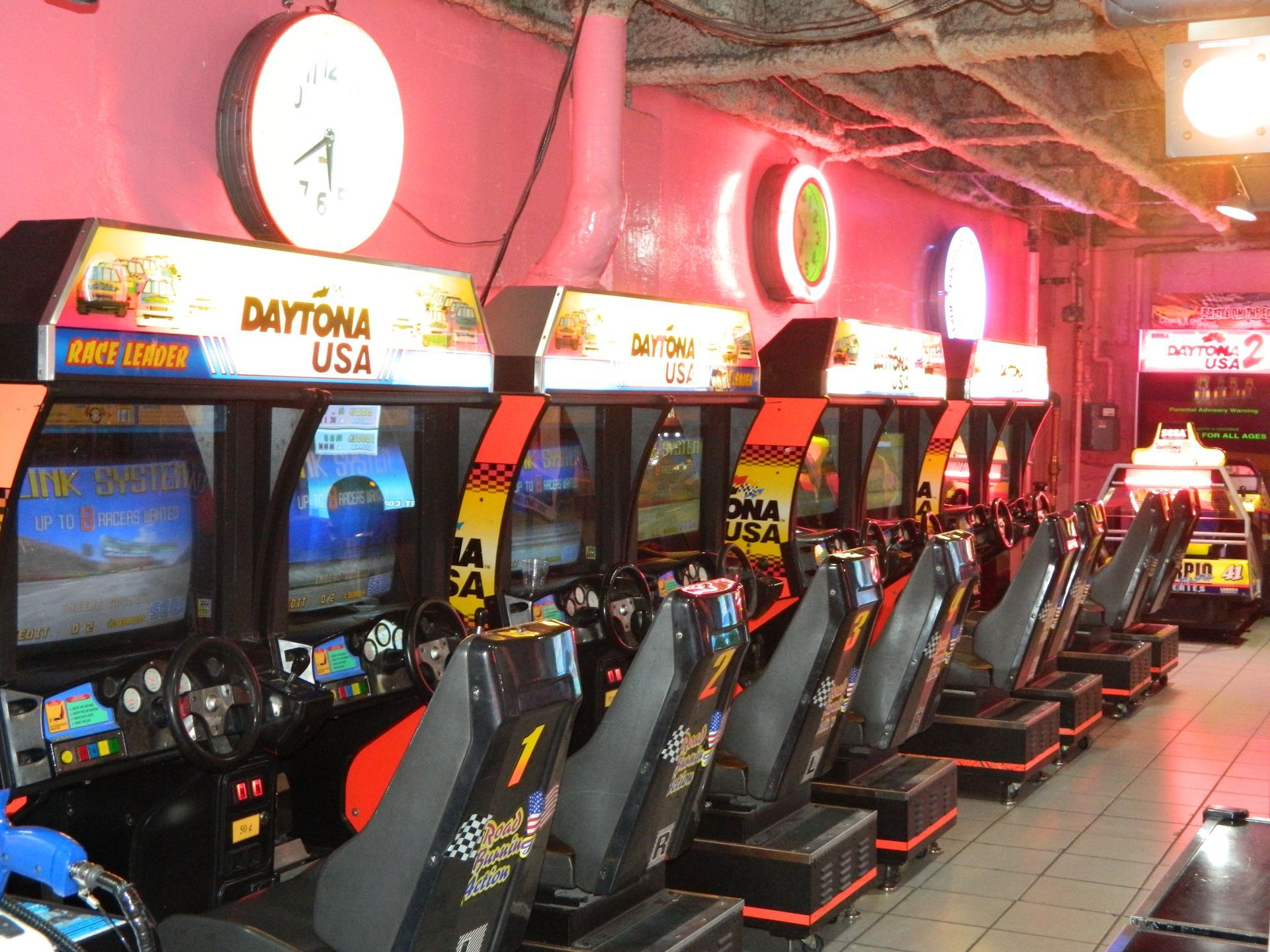 Row of Daytona USA arcade racing game machines in a brightly lit arcade with pink walls and neon lights.