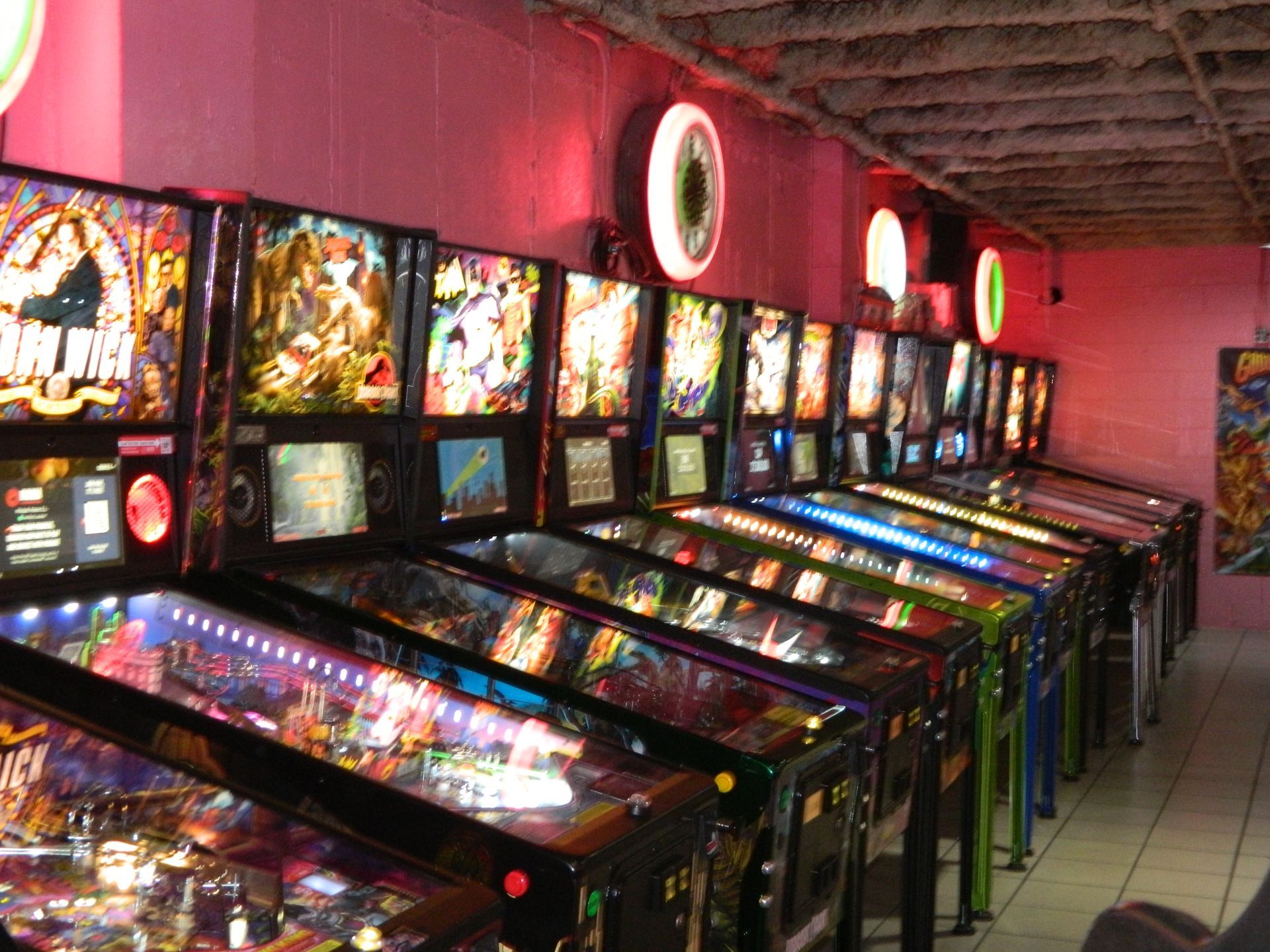 Row of illuminated pinball machines in a dimly lit arcade with pink walls.