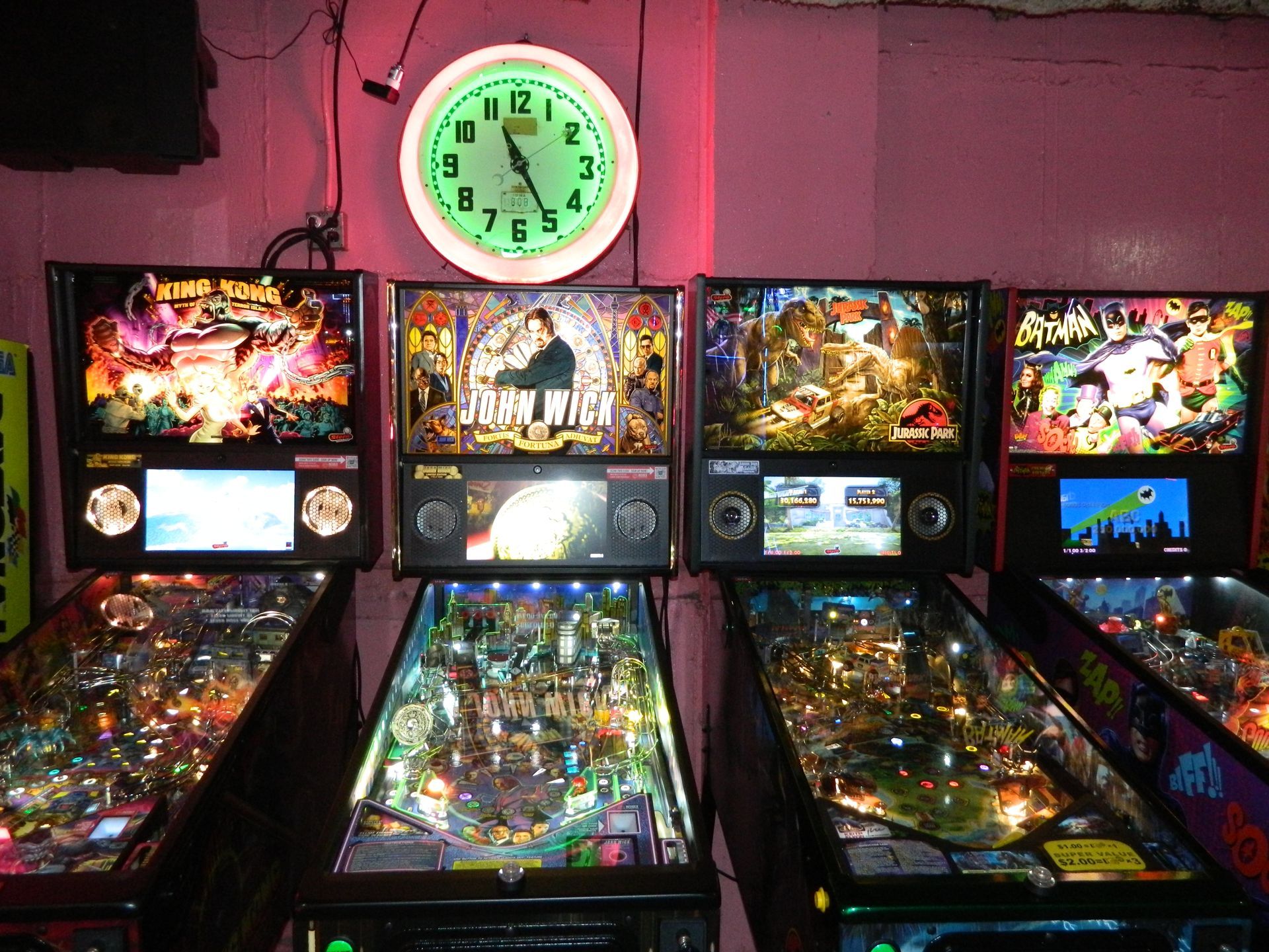 Four pinball machines in a row under a lit clock on a pink wall.