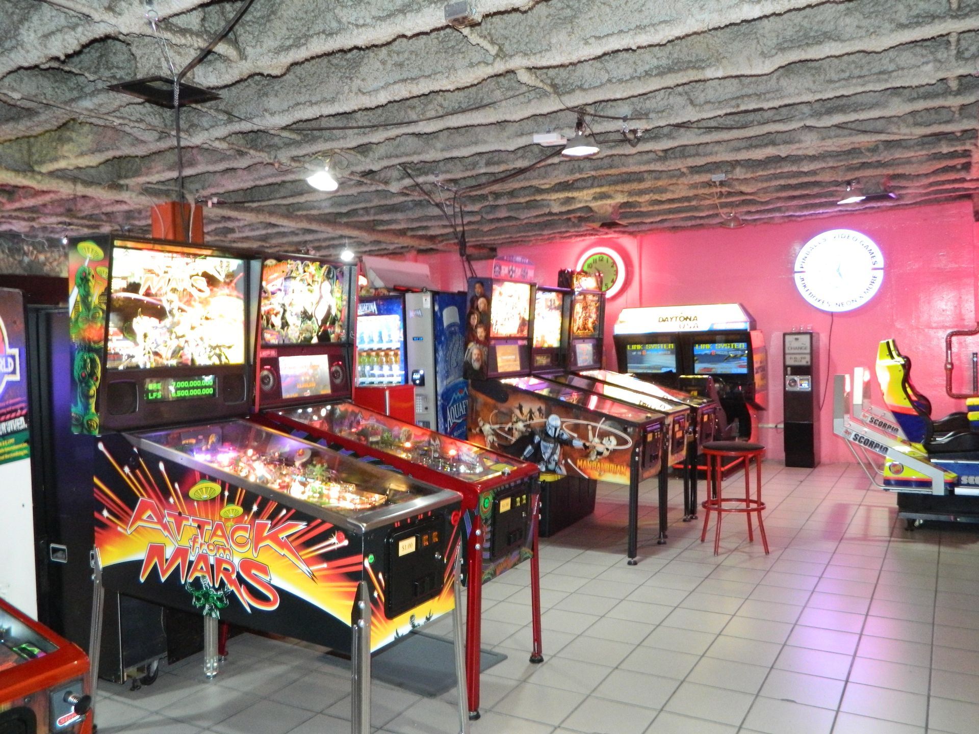 Arcade game room with pinball machines and video games. Pink walls, red stools, white tile floor, and a drop ceiling.