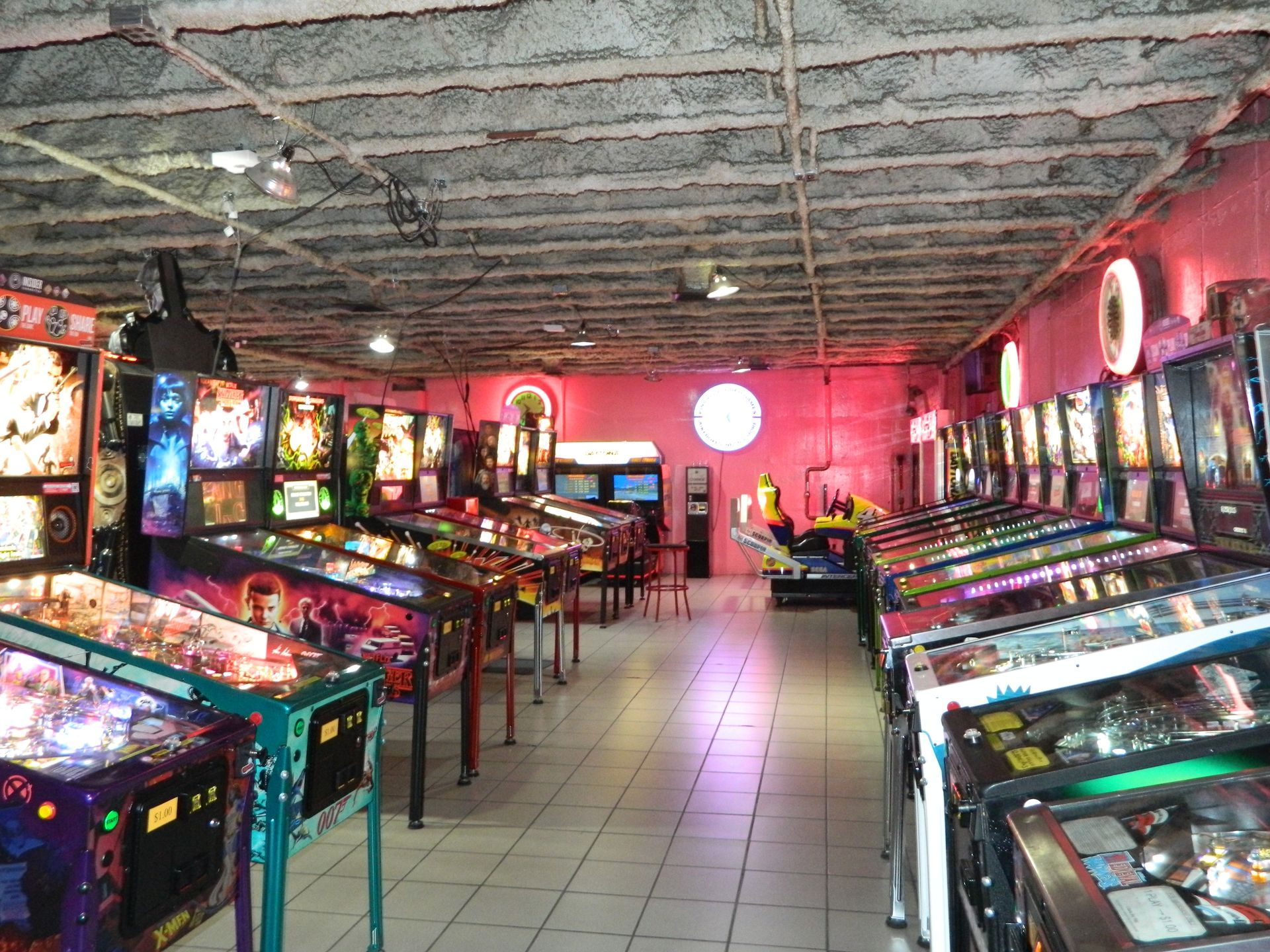 Rows of pinball machines in a dimly lit arcade, pink walls, overhead lighting.
