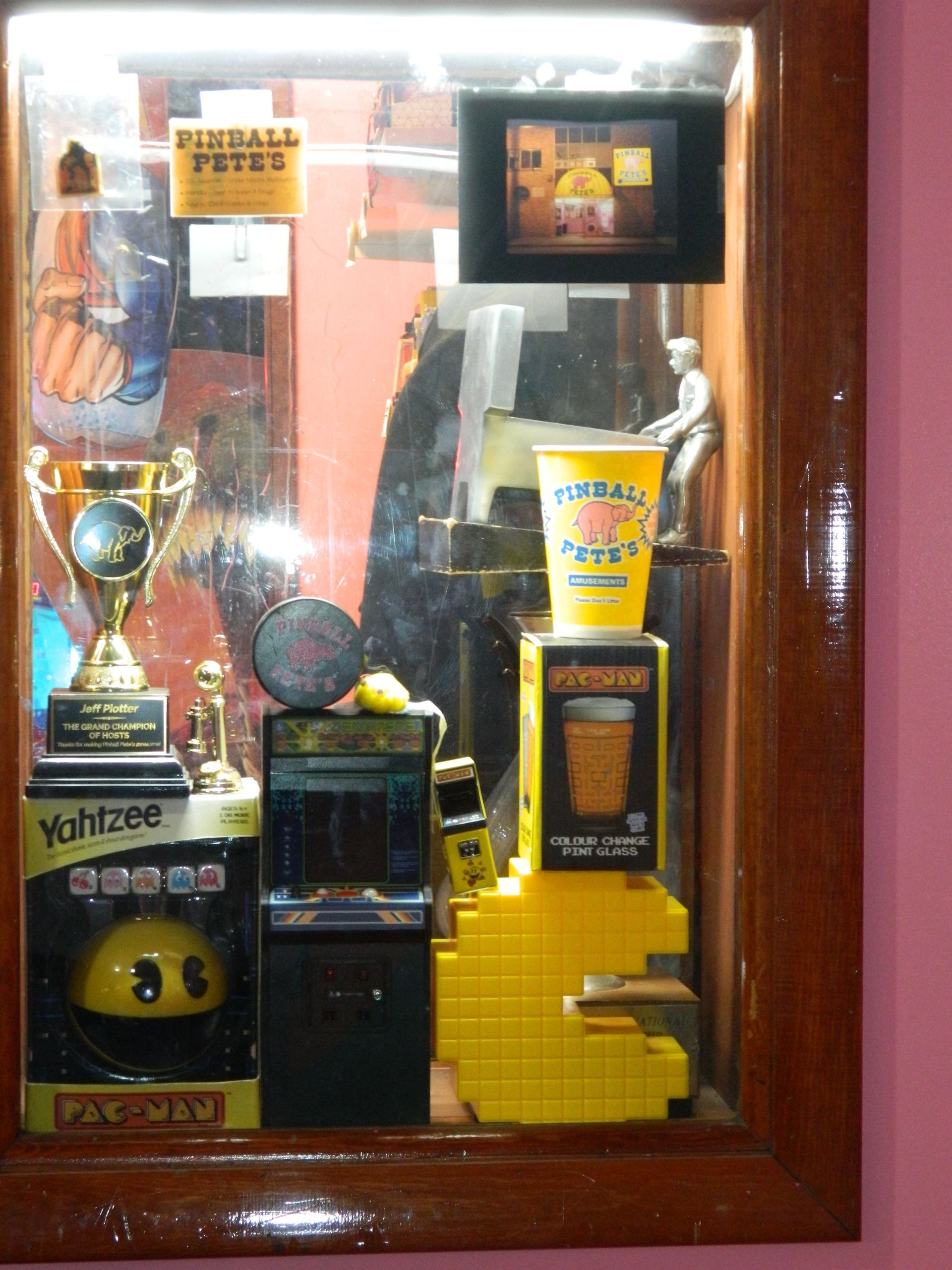 Wooden display case with game-themed items: trophies, Pac-Man, slot machine replica, beer glass, and a small figurine.