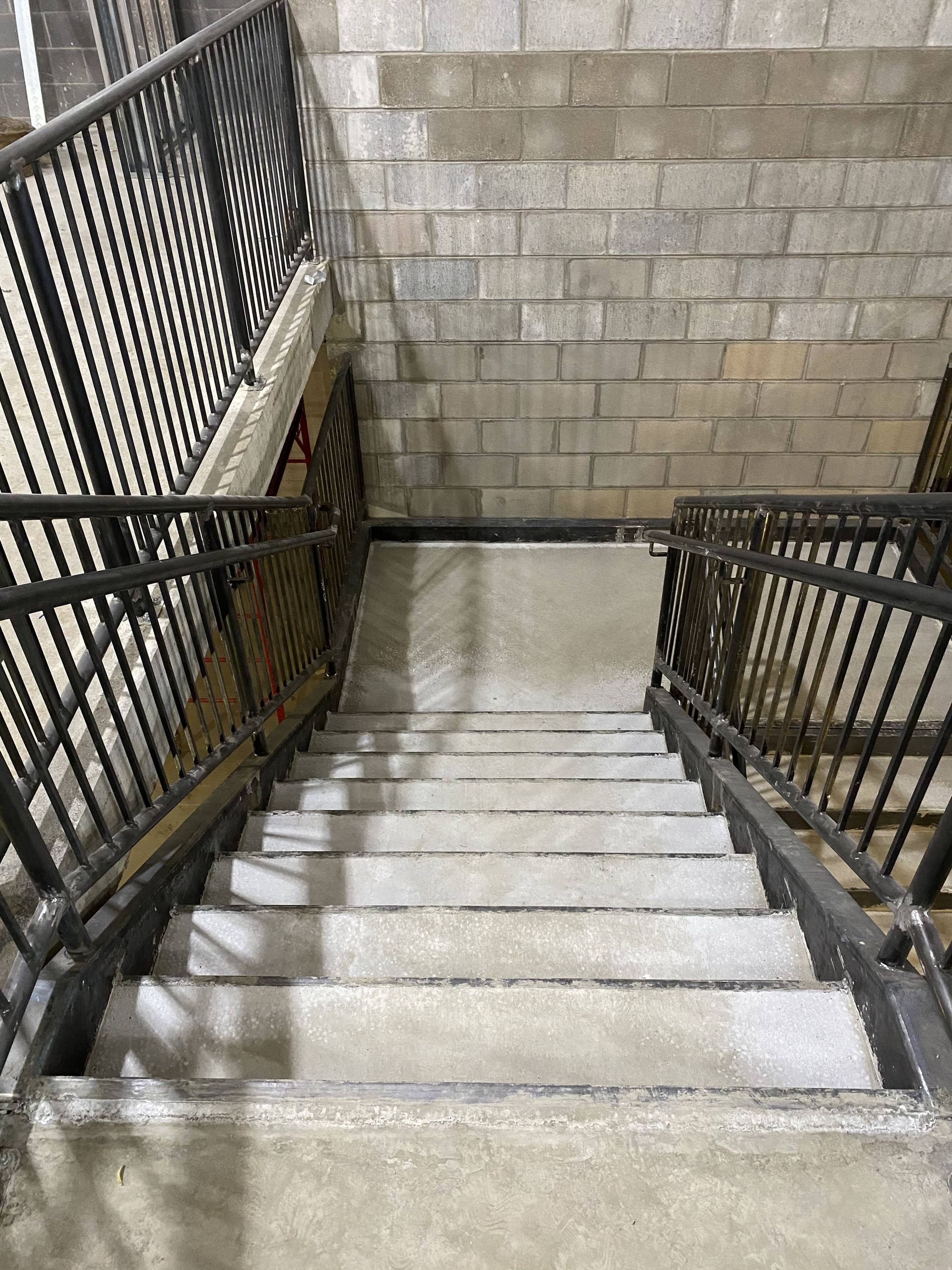 Staircase descends between metal railings, with gray concrete steps and a block wall backdrop.