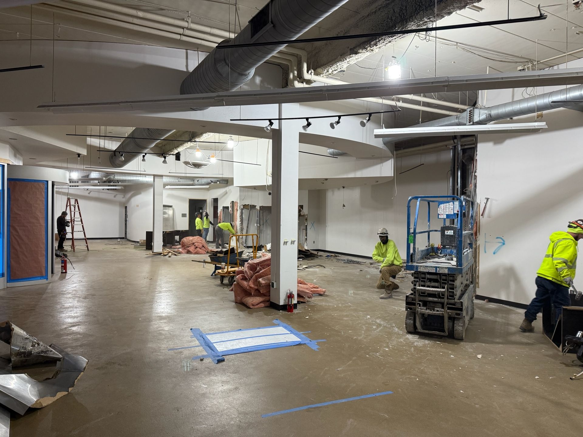 Interior construction site with workers in neon vests, concrete floor, exposed ducts.