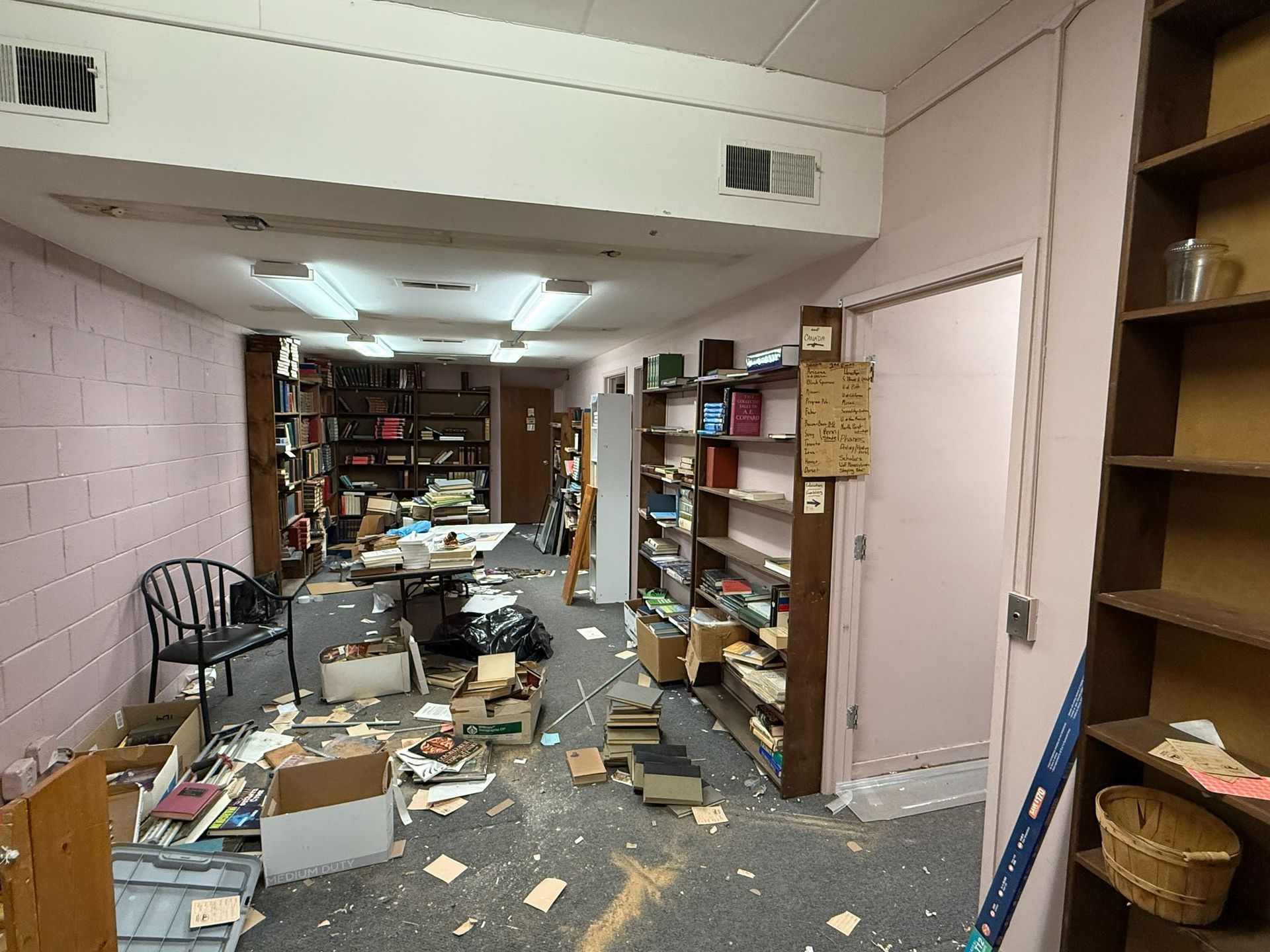 Interior of a cluttered room with bookshelves, debris, and boxes. Pink walls, brown shelves, and a dark chair.
