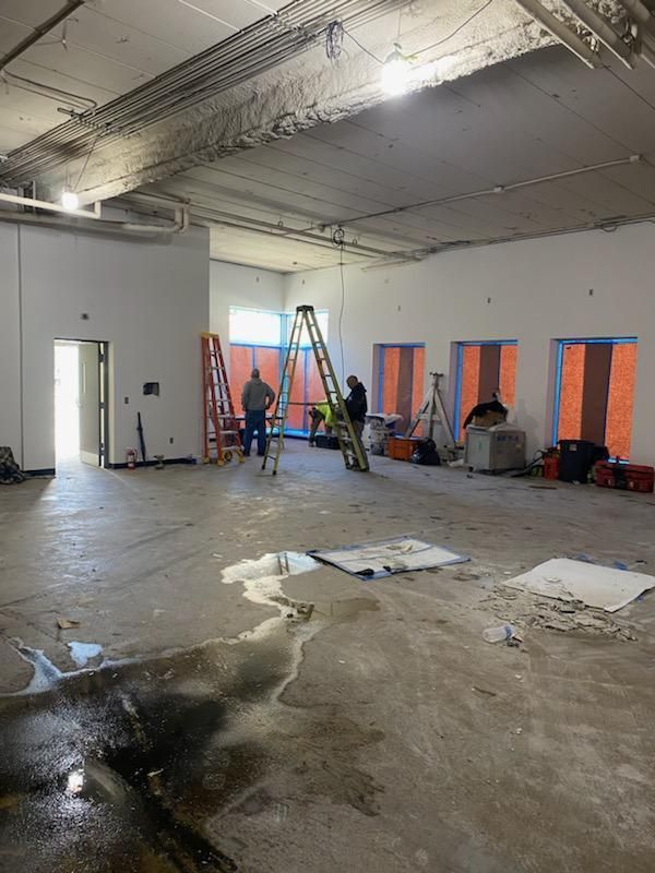 Construction site interior, unfinished walls, concrete floor, workers on ladders, debris scattered.