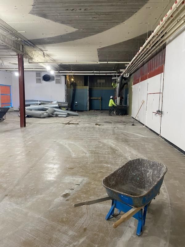 Construction site interior: Wheelbarrow in foreground, workers near center, exposed ceiling, and walls.
