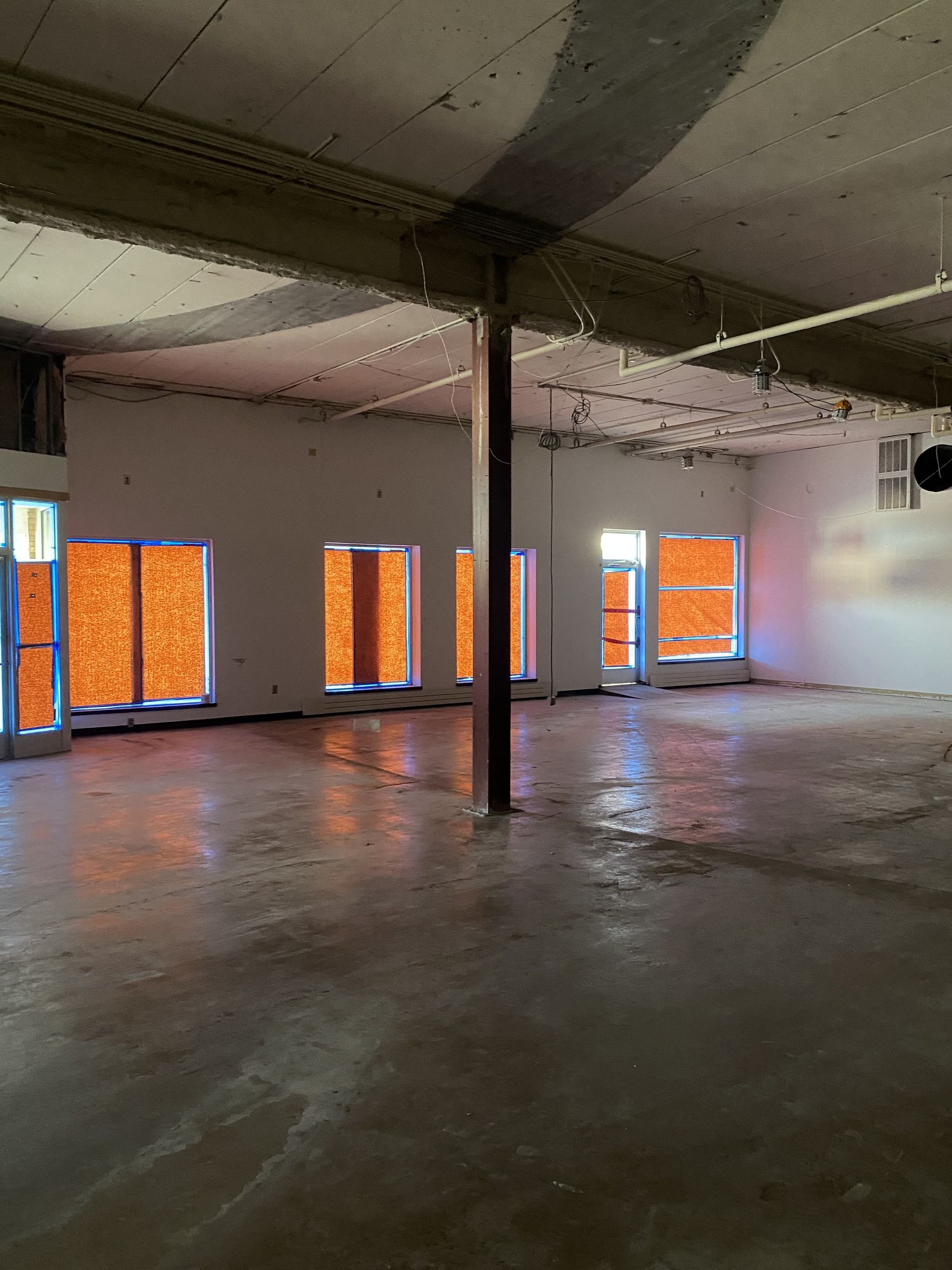 An empty room with concrete floor, windows taped with orange material, and a central support beam.