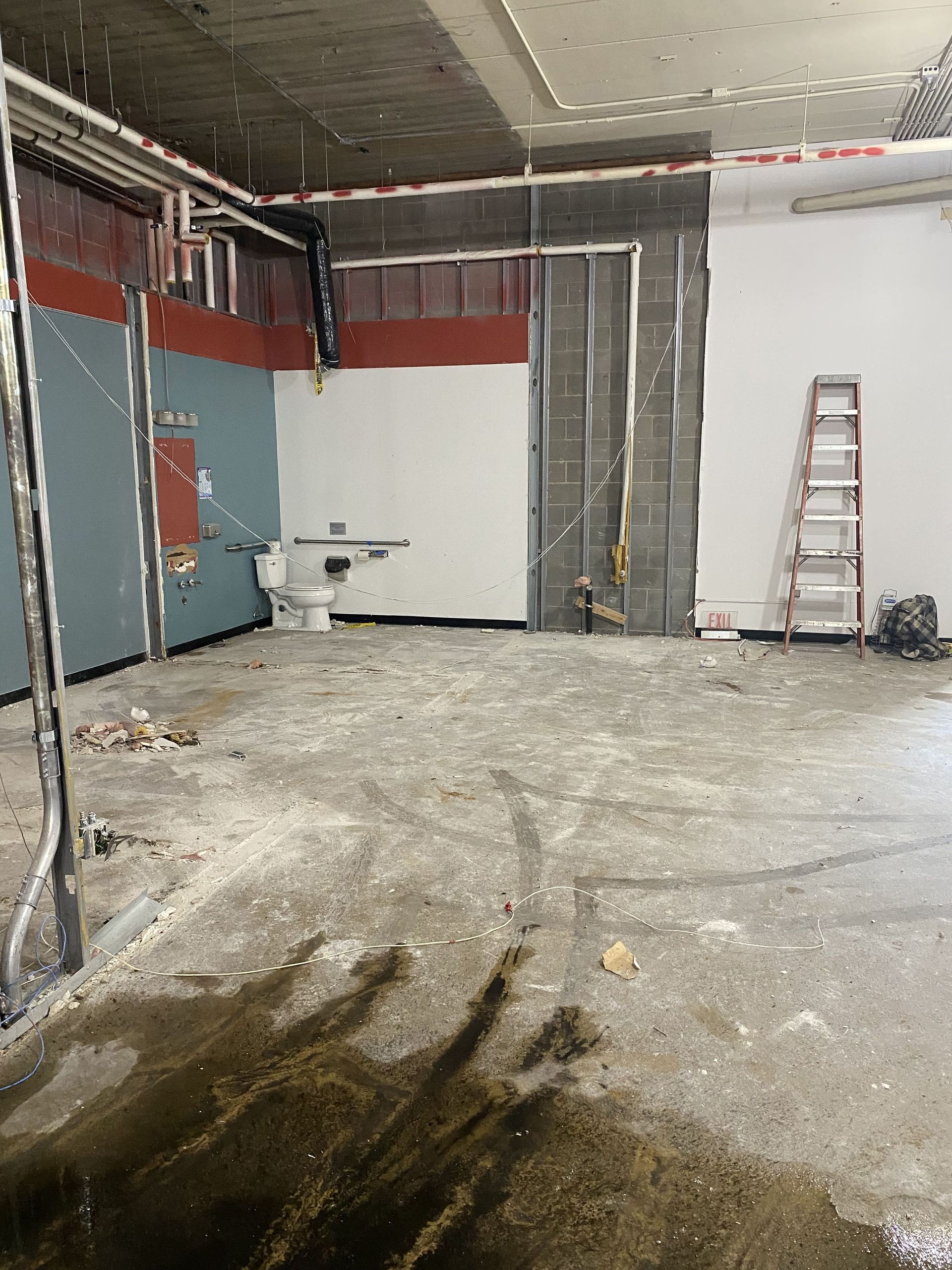 Construction site, interior view. Walls in various colors, exposed pipes, and a ladder are visible. Concrete floor with wet spots.