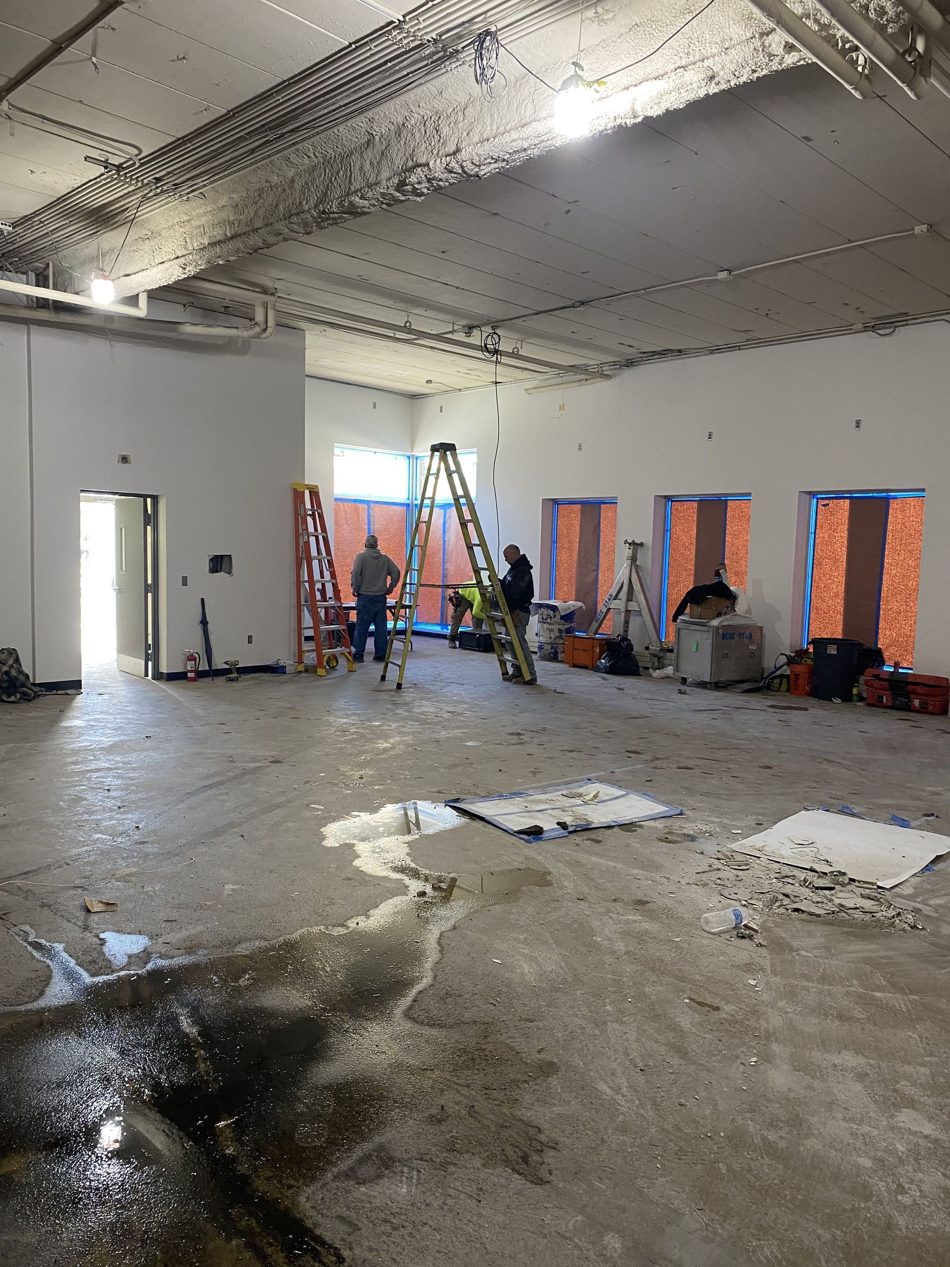 Construction site interior. Workers, ladders, unfinished walls, bare concrete floor, water puddles.