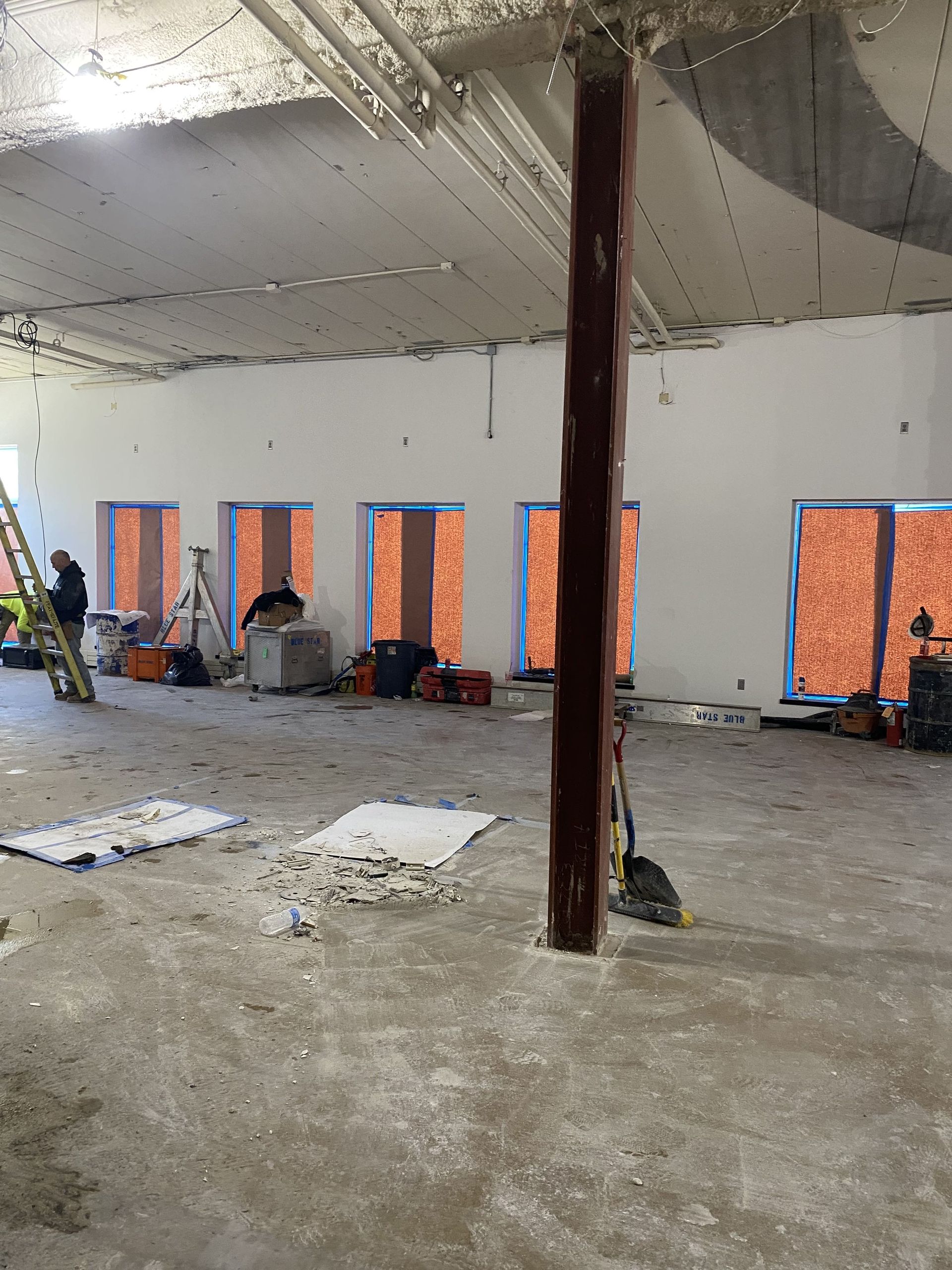 Interior construction site: white walls, exposed ceiling, five boarded-up windows, central column, tools, and a person near a ladder.